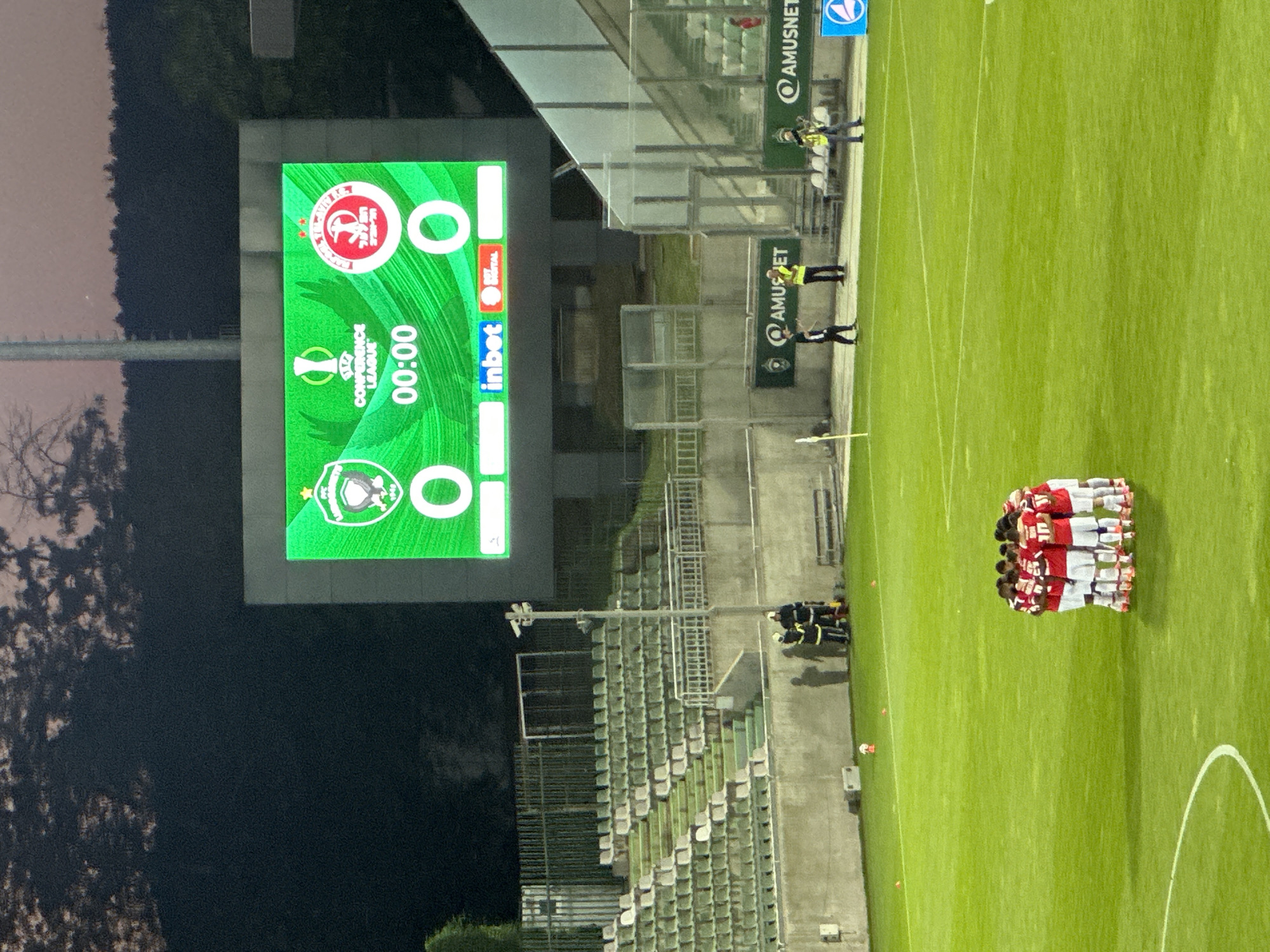 A football (soccer) match is about to begin or has just started, as indicated by the scoreboard displaying "00:00". The image captures a nighttime scene at a football stadium. The scoreboard, a prominent object, shows two team logos and the score. One logo appears to be for a team with red and white as primary colors, and the other has a green and white emblem. The text "CONFERENCE LEAGUE" is visible on the scoreboard, suggesting the match is part of a European competition.
The setting is a football pitch with bright green grass and white markings. In the foreground, a group of players, dressed in red and white kits, are huddled together in a circle, likely for a pre-match or mid-match team talk. To the left of the pitch, several individuals wearing yellow vests and dark clothing are standing near some stadium infrastructure. These individuals might be stadium staff or security personnel.
In the background, empty stadium seats are visible, suggesting that either the crowd is small or the image was taken before the majority of spectators arrived or after they left. The lighting indicates it is nighttime, likely after sunset, and the sky is dark. Some trees and stadium lights are also visible in the periphery. The overall atmosphere appears to be one of anticipation for the sporting event. The text "AMUSET" is visible on banners and signs within the stadium complex, likely indicating a sponsor or the stadium's name.