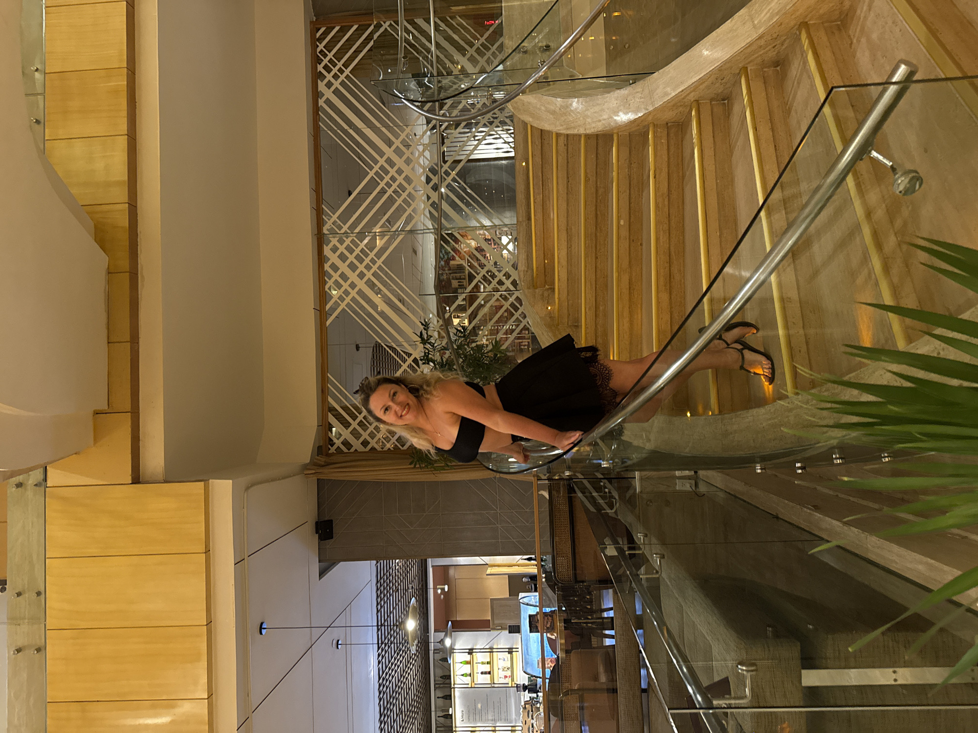 A medium, eye-level shot shows a blonde woman in a black two-piece outfit with a skirt that ends above the knee, posed on the glass-enclosed stairwell of a restaurant. The woman smiles toward the camera. The restaurant interior features wooden details and linear patterns with white accents. A partial view through the glass shows a bar area with displayed bottles and chairs in the background. The scene suggests a modern dining establishment, and the location can be identified as Nha Trang Ward, Vietnam, based on contextual clues and restaurant layout.