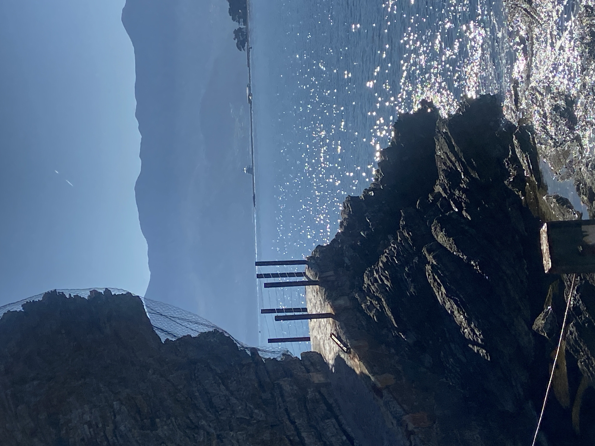 The image depicts a coastline near Budva, Montenegro. The foreground features dark, rocky cliffs with visible geological strata and a safety net secured to the top of one cliff. A structure with protruding, evenly spaced horizontal supports is anchored into the rock face, possibly for engineering or safety purposes. Beyond the rocks is the Adriatic Sea, sparkling with sunlight and reflecting a bright sheen. In the distance, a strip of land or coastline is faintly visible, along with a faint mountainous skyline. The sky is clear and light blue.