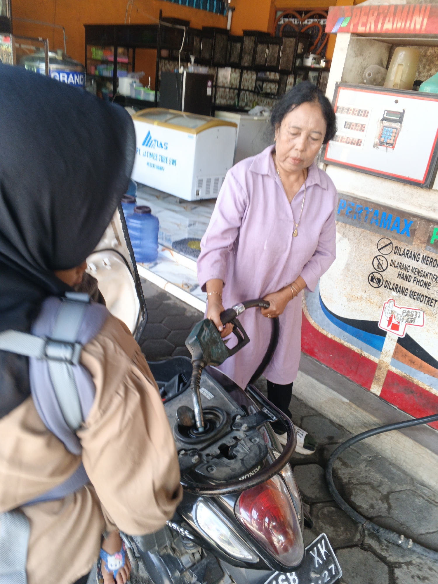 An Indonesian woman, wearing a light purple collared shirt and black leggings, dispenses fuel from a gas pump nozzle into a motorcycle. She is standing next to a scooter that a person wearing a black hijab and brown outer garment is seated on. The gas station appears to be a small, local establishment. Signage visible includes "PERTAMAX" and several warnings in Indonesian, such as "DILARANG MEROKOK" (No Smoking) and "DILARANG MENGAKTIFKAN HAND PHONE" (No Activating Hand Phone). Behind the woman, a white refrigerator unit with the brand "TIAS" is visible, along with other shelves and products within a small store area. The ground is paved with hexagonal tiles. The overall scene suggests a typical roadside gas station in Indonesia, potentially in the city of Padang, given the Indonesian language on the signage.