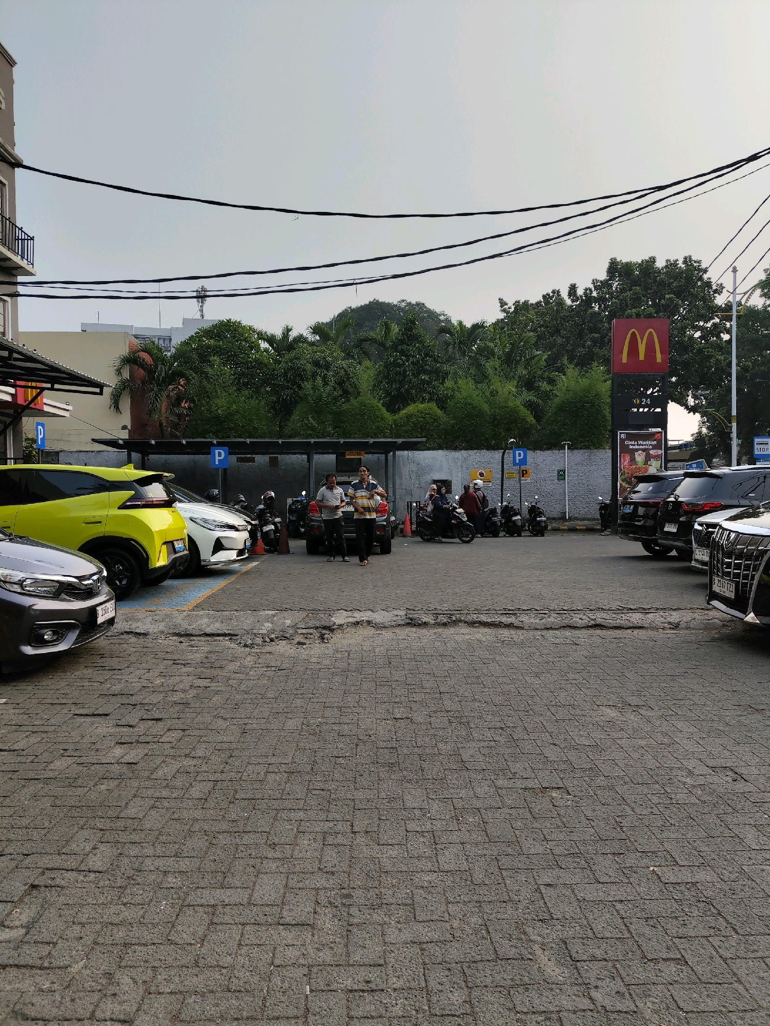 This is a photograph of a McDonald's restaurant in South Jakarta, Indonesia. The image captures the exterior of the restaurant, including its iconic Golden Arches sign, along with a parking lot filled with various vehicles, including cars and motorcycles. In the foreground, several cars are parked, with a yellow SUV prominently displayed on the left. In the middle ground, a group of people are seen standing near parked motorcycles. Behind them, the McDonald's building is visible, and further in the background, lush green trees form a natural backdrop. The sky above is overcast, suggesting a cloudy day. The parking lot is paved with brick-like interlocking concrete blocks.