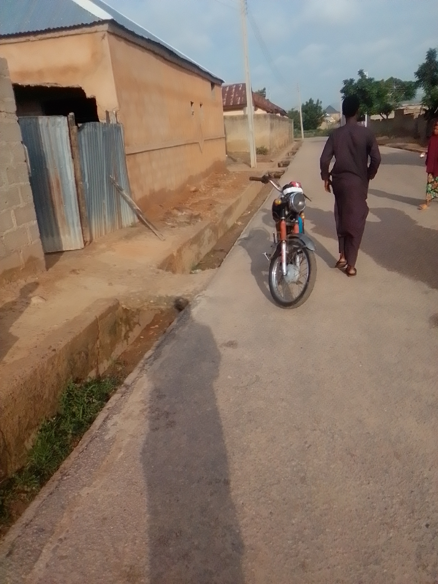 An outdoor, low-angle, eye-level shot shows a dirt road in the city of Liji Hill, Nigeria. In the foreground, a motorcycle is parked on the right side of the road. The motorcycle is red and silver and has a single headlight. Behind the motorcycle, a man wearing a dark brown robe walks away from the camera. To the far right, a woman in a red top and floral skirt walks on the sidewalk. The background shows a building with a corrugated metal roof and a concrete wall. The sky is overcast.