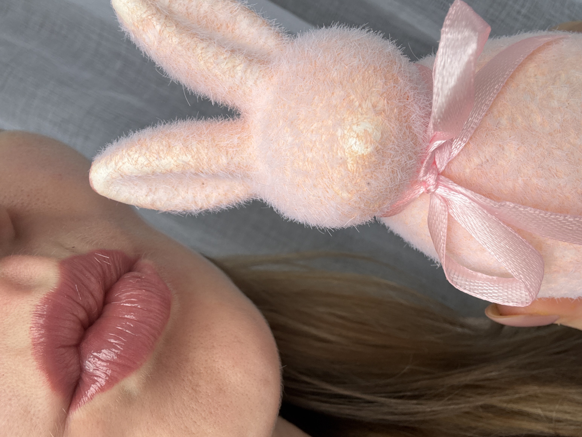 A close-up shot shows a person's face juxtaposed with a pink, fluffy bunny toy tied with a pink ribbon. The person's face is positioned on the left side of the frame, featuring prominently glossy, pink-toned lips. The bunny toy is placed on the right, with its ears extending towards the upper left corner. The background is a soft, blurry grey-white, possibly fabric. Hair is partially visible near the face, appearing light brown. The location is not discernible from the provided image.