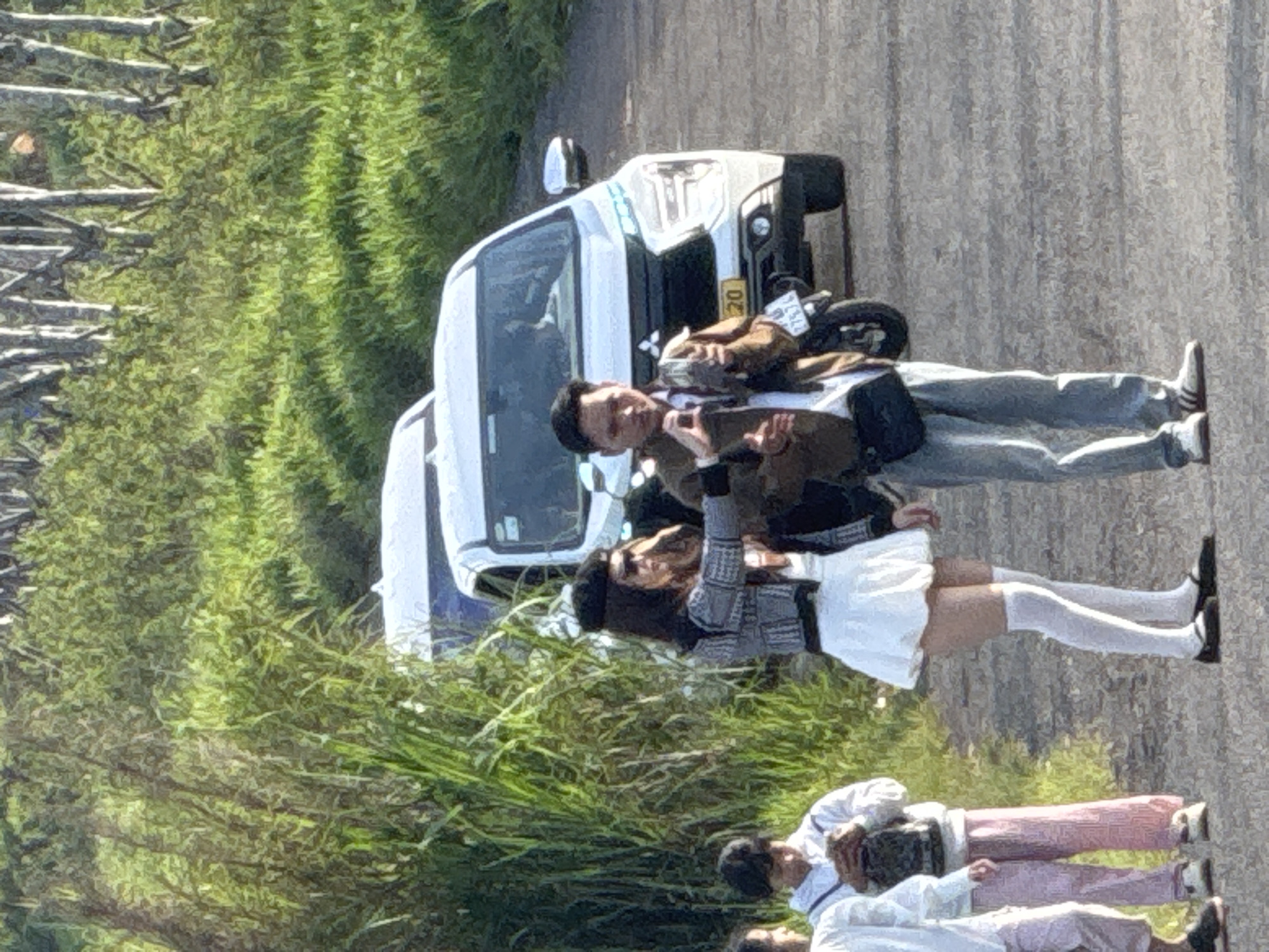 This media file captures an outdoor scene in Trường Thọ, Vietnam, featuring several individuals and a white van. The image appears to be taken during daylight, with bright sunlight casting shadows and illuminating the environment.
In the foreground, a young woman is wearing a plaid jacket, a white skirt, and white knee-high socks, holding a bag. Next to her, a man in a brown jacket is standing and appears to be gesturing. Behind them, another person is visible, dressed in a white top and light pink pants. Further in the background, part of a white van is visible, with a license plate that reads "20" and some unreadable characters. Another person is seated on a motorcycle, also partially visible.
The setting includes a paved road and a grassy area with trees or bushes. The overall atmosphere suggests a casual outing or a stop during a journey. The individuals seem engaged in conversation or observation, with expressions that are difficult to discern clearly due to the image quality. The image is oriented vertically, with the subjects appearing sideways.