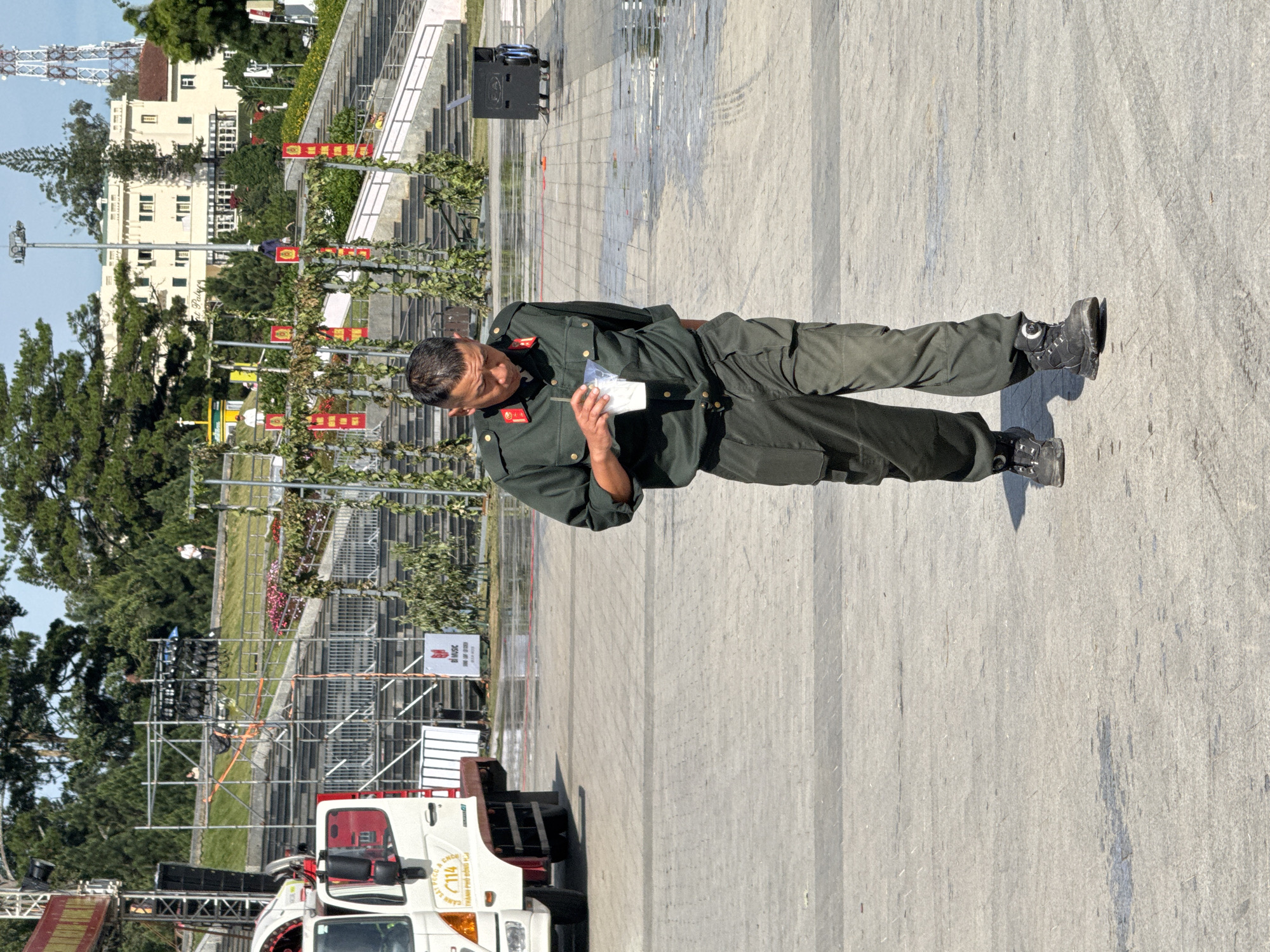 A man in a dark green uniform walks across a paved plaza, holding a clear plastic cup with a white beverage. The uniform consists of a button-up shirt with epaulets and red trim on the collar, and loose-fitting cargo pants. He wears dark, sturdy boots. The plaza is wet in places, indicating recent rain or cleaning. In the background, a white truck with a red cab is parked to the left. Further back, a tiered seating area with metal scaffolding and lighting equipment is visible, suggesting an outdoor event space. Beyond the seating, trees and a light-colored building with multiple windows and a pitched roof are prominent. A cell tower stands tall in the distance. Red banners with Vietnamese writing hang from trellises in front of the seating area. The scene appears to be a public space in Da Lat, Vietnam, likely a plaza or park designed for events, given the staging and seating.