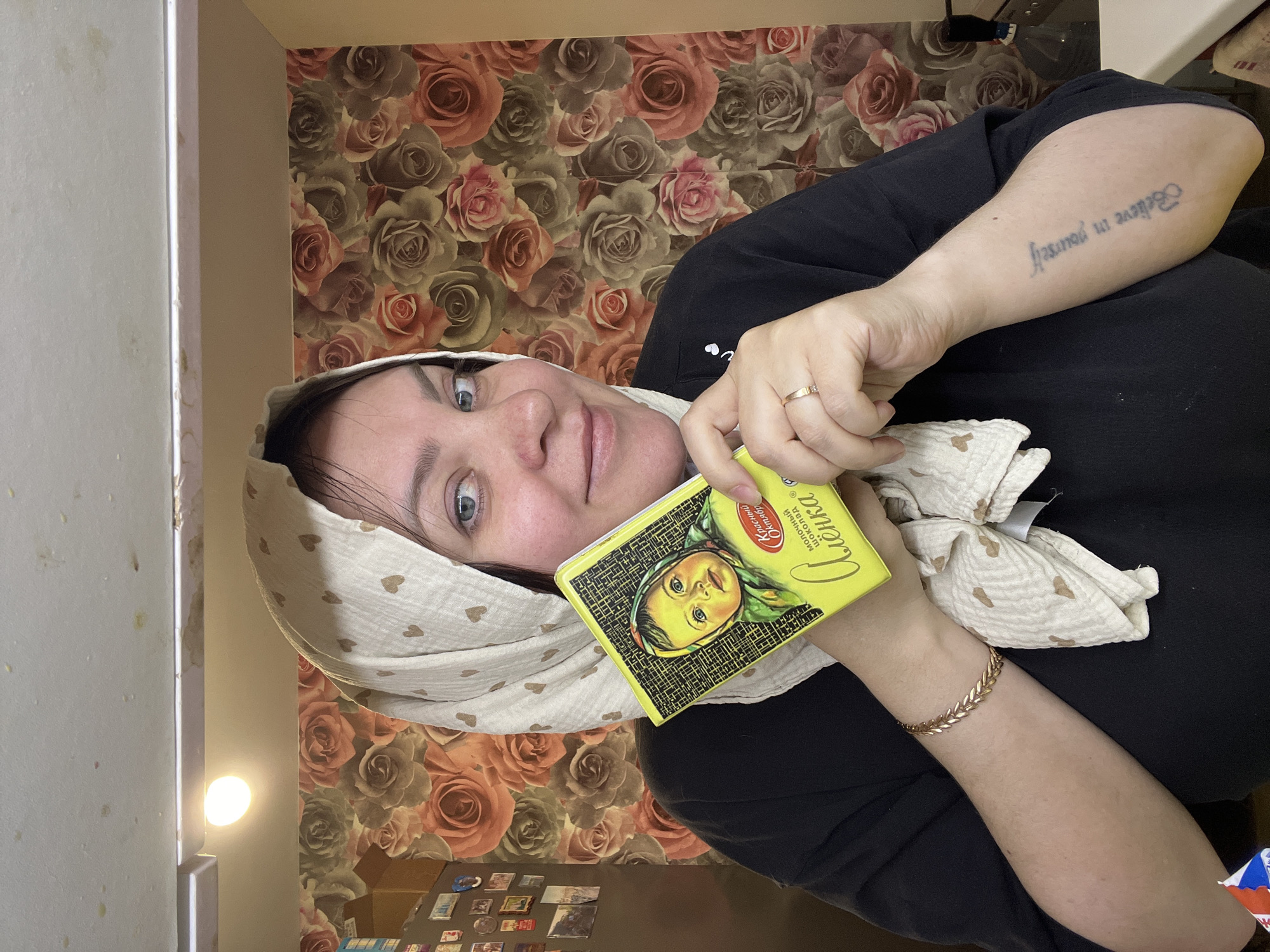 A person, likely female, is captured in a close-up, upward-angled selfie, set indoors in Noviy, Russia. She is smiling slightly and looking towards the camera, holding a yellow rectangular box with both hands. She wears a plain black top and a light-colored head covering, possibly a hijab or scarf, adorned with small light brown heart patterns. A gold-colored ring is visible on her right ring finger, and a gold-colored bracelet with a leaf-like design is on her left wrist. On her left forearm, a tattoo reads "Believe in yourself" in a stylized script.
The background features a prominent floral wallpaper with large, muted pink and gray rose patterns. To the left, a portion of a vertical white surface, possibly a refrigerator or cabinet door, is visible, showing some minor smudges. A bright, circular light source is present in the upper left background, casting a soft glow.
The yellow box she is holding displays a distinctive image of a young baby or child, recognized as the mascot for a popular confectionery brand. Visible Russian text on the box includes "Алёнка" (Alyonka) in large red script, with "Красный Октябрь" (Krasny Oktyabr, or Red October) and "молочный шоколад" (milk chocolate) also present. The overall scene suggests a casual, personal moment.