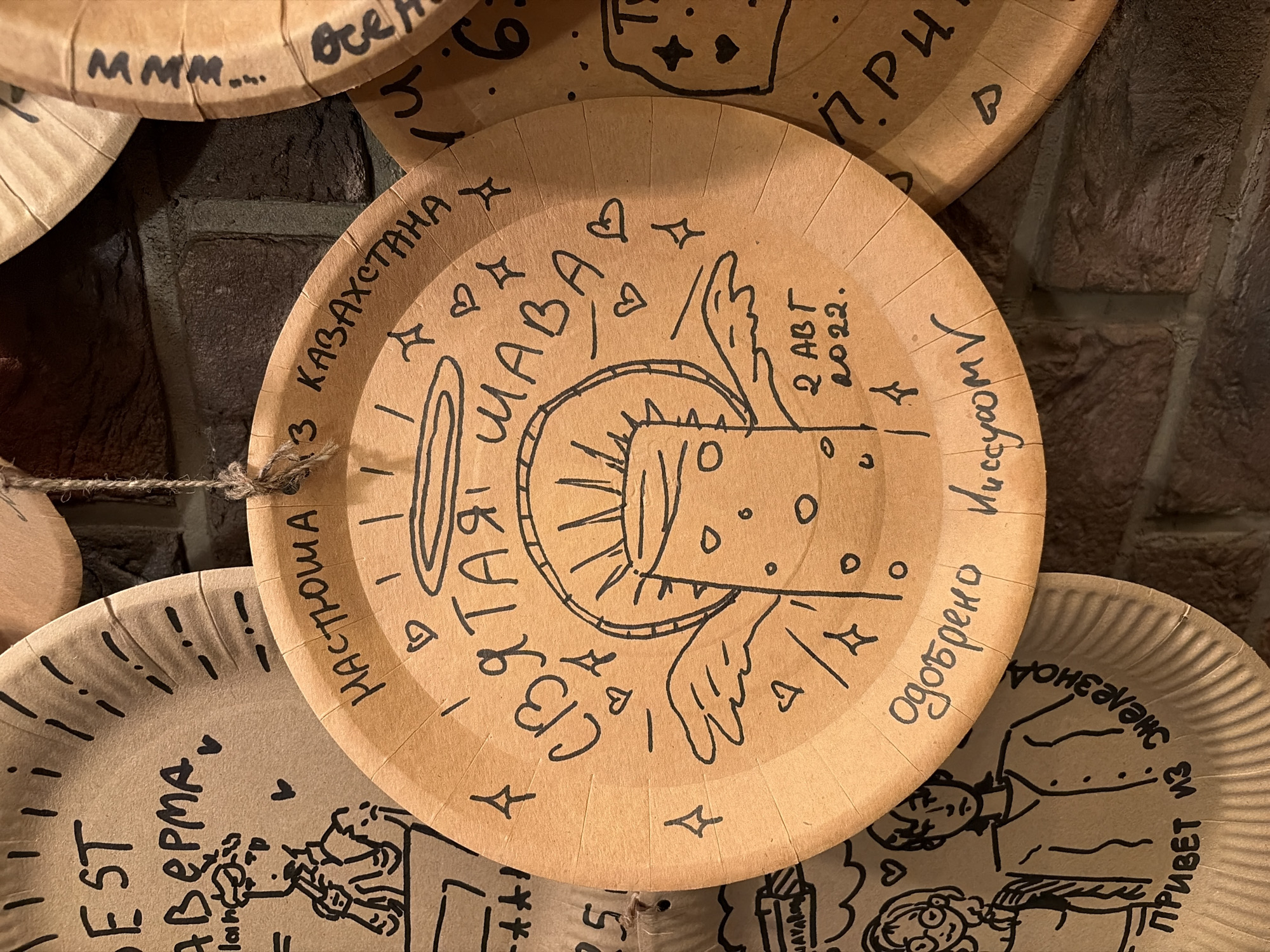 The image shows a close-up of several paper plates with drawings and handwritten text, possibly displayed as decorations. The plates are brown and appear to be slightly wrinkled.
The central plate features a drawing of a mushroom-like figure with wings and a halo, suggesting an angelic or spiritual representation. Surrounding this figure are handwritten words in Russian: "НАСТЮША" (Nastyusha) at the top, and "СВЯТАЯ ШАВА" (Holy Shava) arching over the drawing. Below the drawing, the date "2 АВТ 2022." (August 2, 2022) is written, along with "одобрено ЛисууомV" (Approved by LisuumV). Various decorative elements like small hearts and stars are scattered around the drawing.
To the left of the central plate, another plate is partially visible with a drawing of two people and some text. One person is depicted wearing glasses and holding something, while the other stands with a stern expression. The text "ПРИВЕТ ИЗ ЖЕЛЕЗНОД" (Greetings from Zhelezno D) is written at the bottom of this plate. Above this, the word "BEST" and "АВЕРМА" are visible, along with a drawing of a person with a pipe.
To the right of the central plate, another plate has the text "МММ..." (Mmm...) and "все" (all) written on it. Further to the right, parts of other plates are visible with minimal text and drawings.
The plates are hanging against a dark brick wall, and a piece of twine is visible, suggesting they are suspended. The overall impression is that of a whimsical or artistic display of personal creations or souvenirs. The image is taken from a close-up perspective, with a shallow depth of field blurring the background. The lighting appears to be ambient, casting soft shadows.
