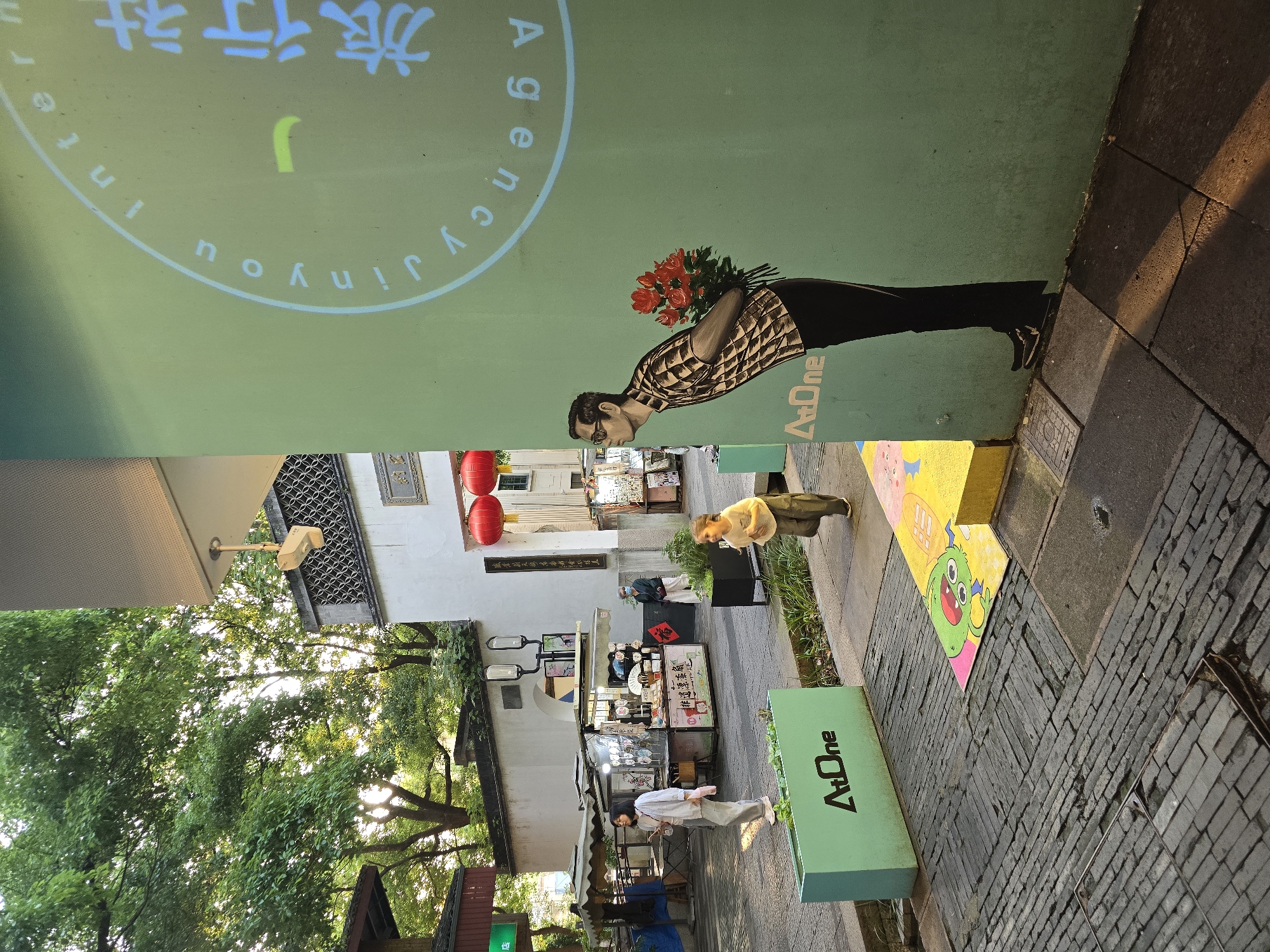 The image captures a street scene in Shangcheng District, China, with a mural of a man holding flowers as a prominent element. The mural is painted on a light green wall and depicts the man from the waist down, with his back turned, concealing a bouquet of red flowers behind his back. Above the mural, a circular graphic with text in both English and Chinese is projected or painted, with the English text partially visible as "Agency jinyou Intern".
In the foreground, a young child with blonde hair, wearing a light-colored t-shirt and dark pants, is in motion, appearing to run or jump across the paved area. The ground is a mixture of dark brick pavers and some lighter concrete slabs. A vibrant, cartoon-like mat featuring a green monster is laid out on a yellow and pink base. To the left of the child, a light green planter box with the "AtOne" logo is visible.
Further back in the scene, there are food stalls or market kiosks, with awnings providing shade. Two women are walking in the background. One woman in a light-colored top and pants is seen from the side, and another woman in darker clothing is partially visible near a stall. Red lanterns are hanging from a traditional-style building entrance. The environment is outdoors, with trees and foliage visible in the upper left portion of the image, suggesting a pleasant, possibly warm, climate. Sunlight casts distinct shadows across the paved ground.