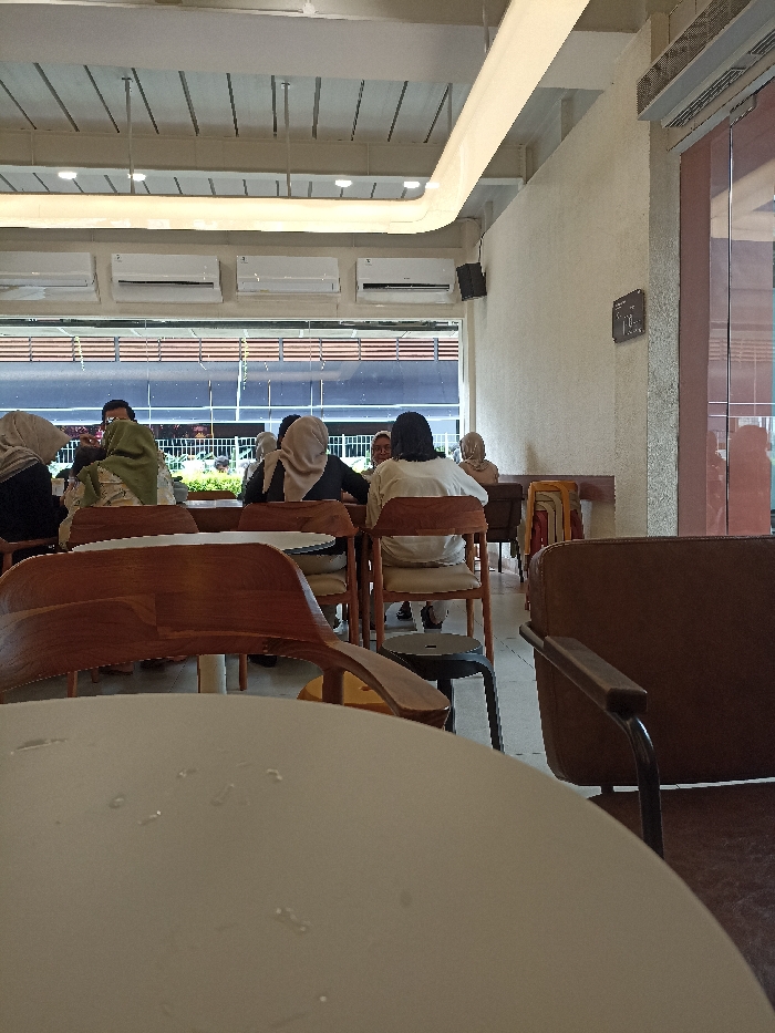 The image shows a cafe interior in Purwokerto, Indonesia, located at latitude -7.42 and longitude 109.245. Six people are seated at tables, with two people in the foreground obscured from view. The people are wearing casual clothing, and some are wearing headscarves. There are tables and chairs throughout the cafe, with a white circular table and a brown wooden chair in the foreground. There is a large window behind the tables, showcasing a view of a building with a tiled roof. The cafe has a white ceiling with recessed lighting, a light brown wall, and tiled floors. A brown door is visible on the right side of the image. The scene is calm and peaceful, with a casual atmosphere.