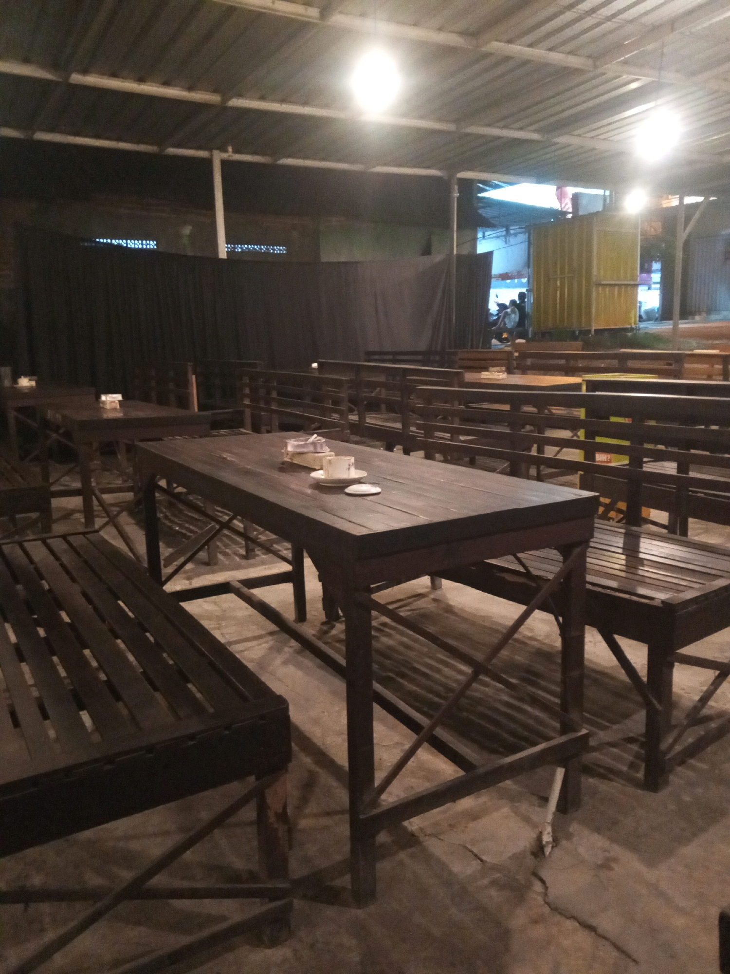 The image depicts an outdoor seating area, likely a restaurant or cafe, with a rustic aesthetic. The furniture consists of dark wooden tables and benches arranged in rows. The tables have a simple, planked design, and some are set with plates and cups. The seating area is covered by a corrugated metal roof supported by metal beams, with exposed lighting fixtures. The ground appears to be concrete, with visible cracks in some areas. In the background, there are glimpses of buildings, a yellow structure, and several people, some appearing to be sitting or standing near motorcycles. The overall lighting suggests it is evening or nighttime. The scene is located in Tambakagung, Indonesia.