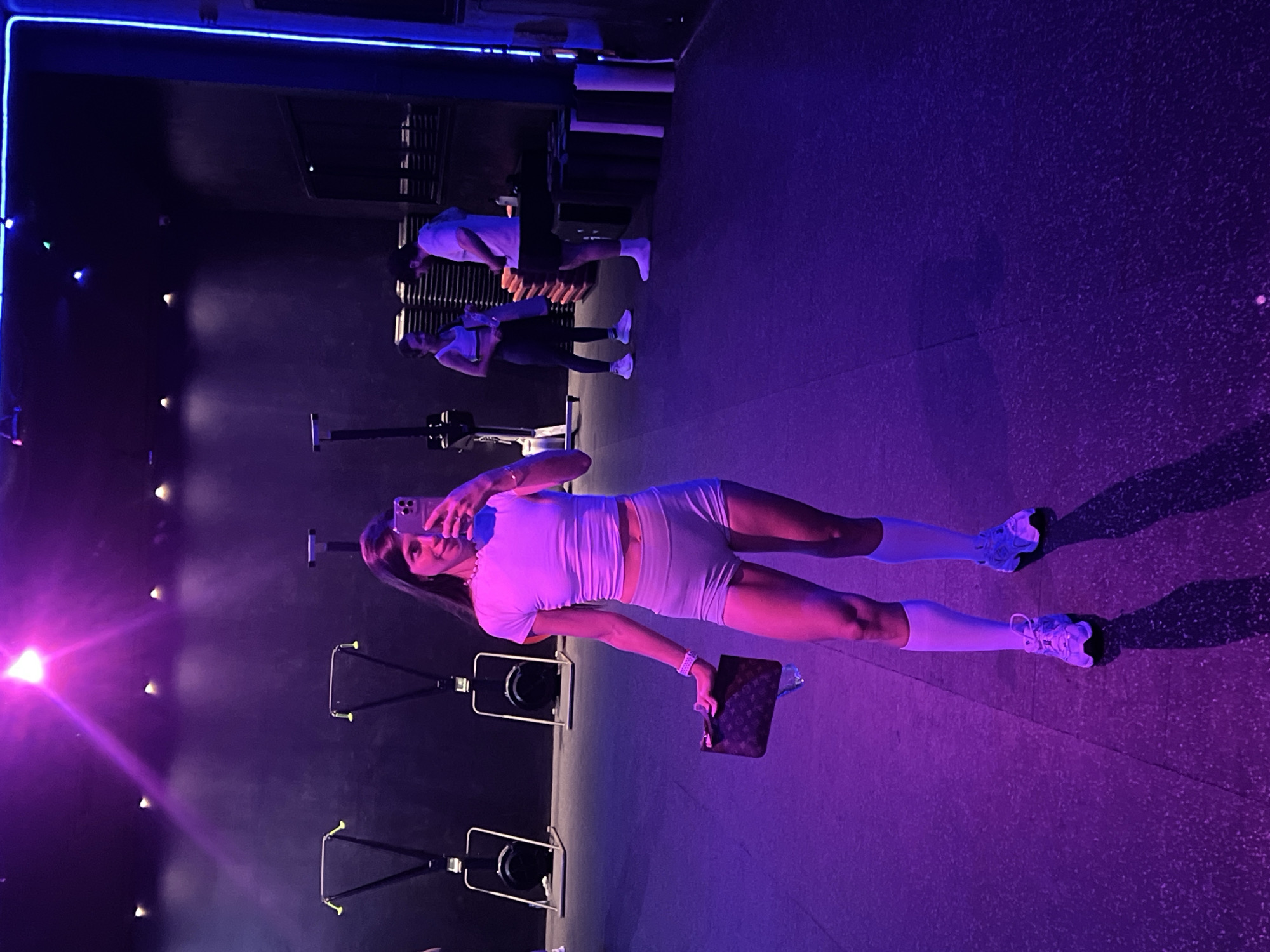 A woman in athletic wear, a crop top and shorts, poses for a selfie in a gym. She holds a clutch in her left hand and a water bottle in her right. The gym is dimly lit with purple and blue lights, and rowing machines are visible in the background. Two other individuals, a man and a woman, are also present in the gym, conversing near a stack of mats. The floor is dark and reflective.
