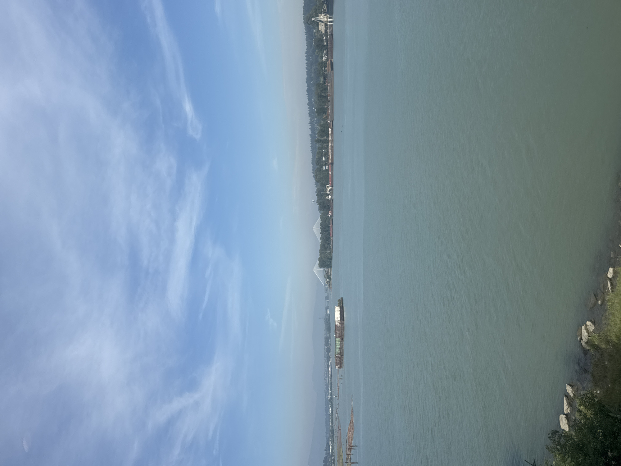 This is a wide shot of the Fraser River in New Westminster, Canada, on a clear day with a few wispy clouds in the sky. The water is a calm, blue-green color. In the middle of the river, a large barge is loaded with what appear to be shipping containers, some of which are green. To the left of the barge, there is a cluster of logs floating in the water. In the distance, a suspension bridge spans the river. The shore on the far side of the river is lined with trees and buildings, suggesting an urban or industrial area. The foreground of the image shows a rocky shoreline with some green vegetation. There are no people visible in the image.