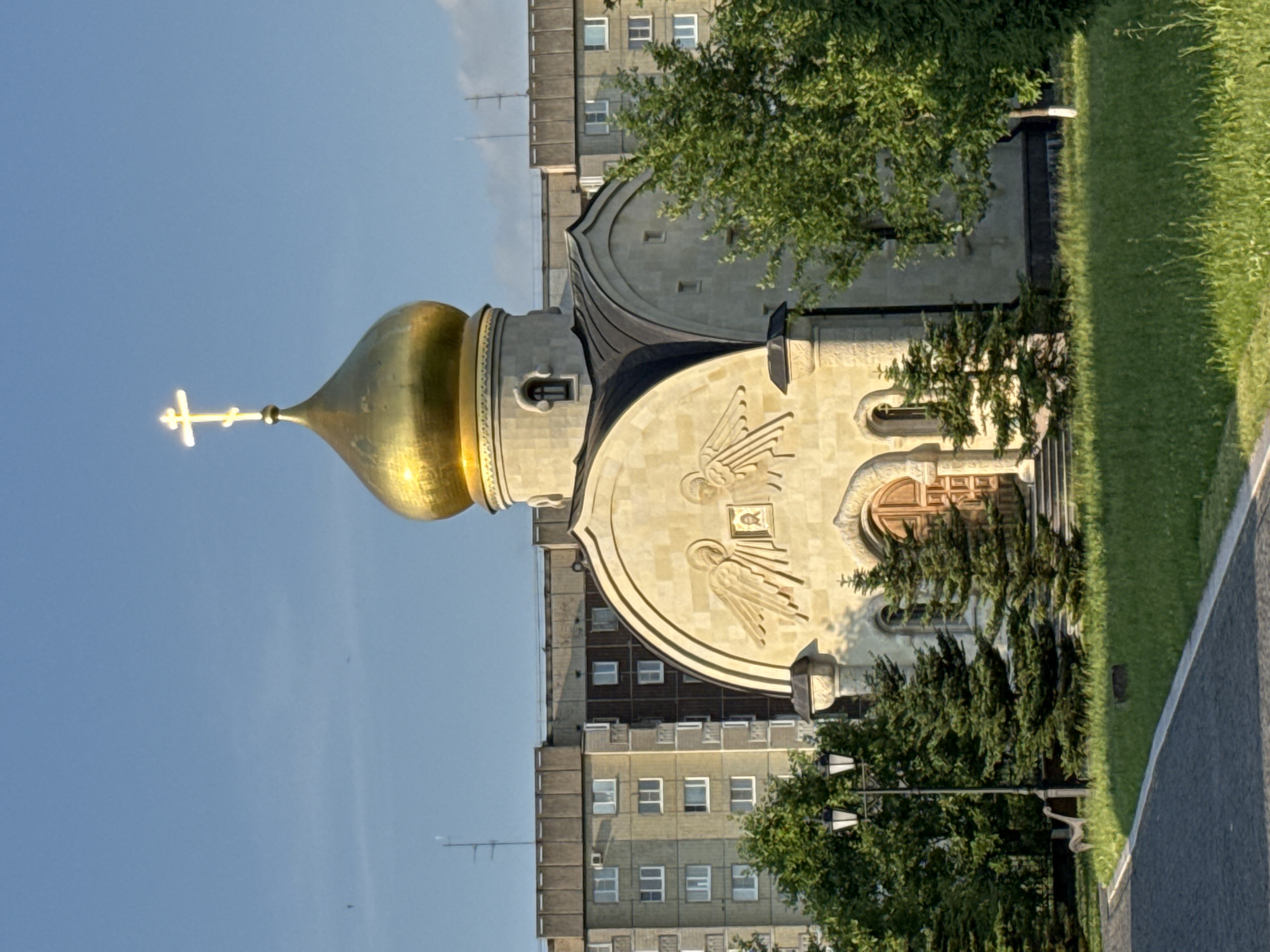 The image shows an Orthodox church with a golden dome and a cross on top, located in the city of Kirishi, Russia. The church is made of light-colored stone and has carved angels above the entrance. It is surrounded by green trees and grass, and in the background, there are tall apartment buildings. A lamppost with two lights stands to the left of the church, and a paved walkway leads towards it. A small, light-colored decorative sculpture resembling a dog is visible near the lamppost. The sky is clear and blue, indicating a sunny day.