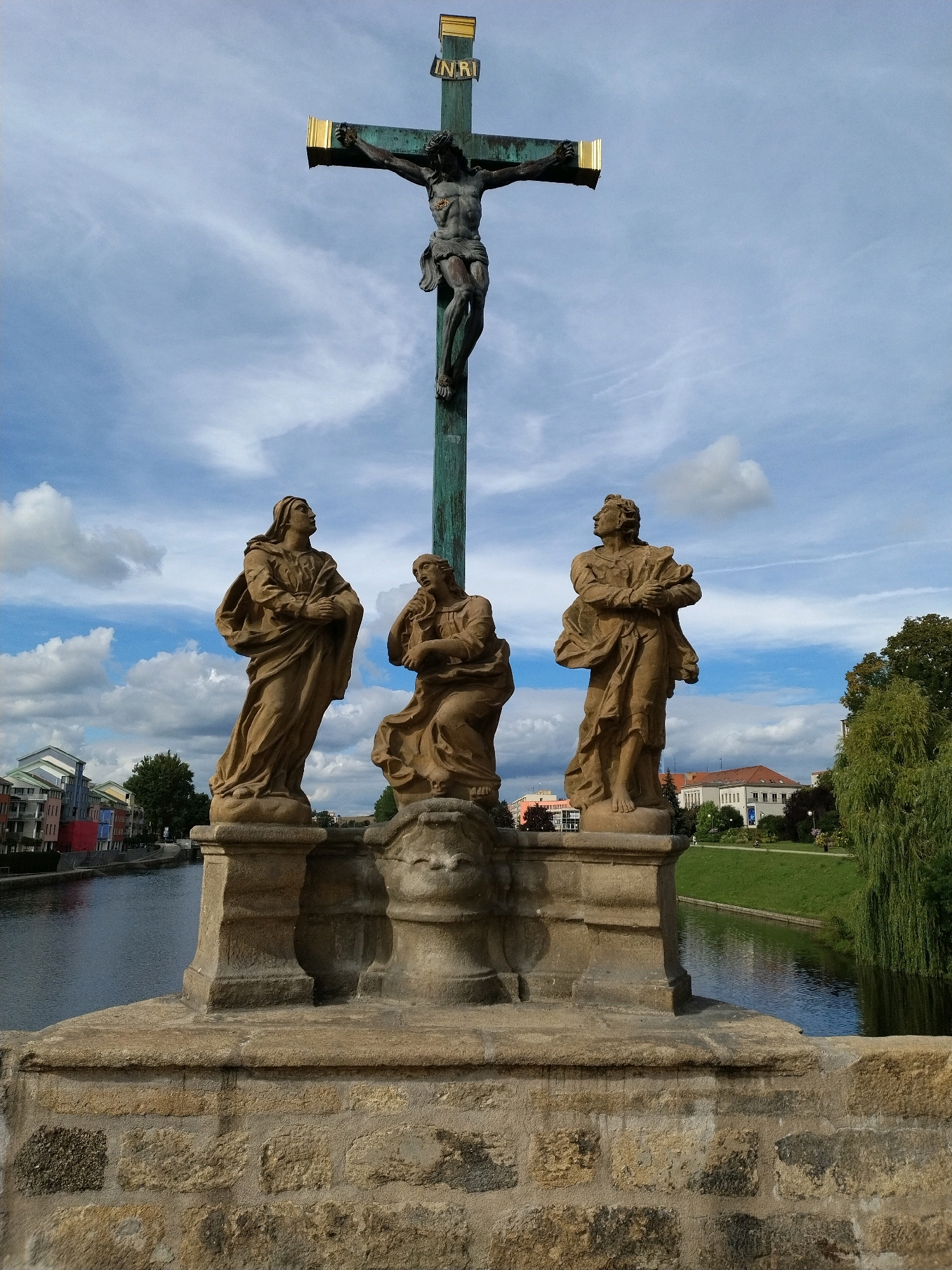 The image shows a statue of Jesus on the cross at the top of a bridge in Písek, Czechia. Below the cross, there are three statues of women. The bridge is made of stone and overlooks a river with buildings on the other side. The sky is blue with some clouds.