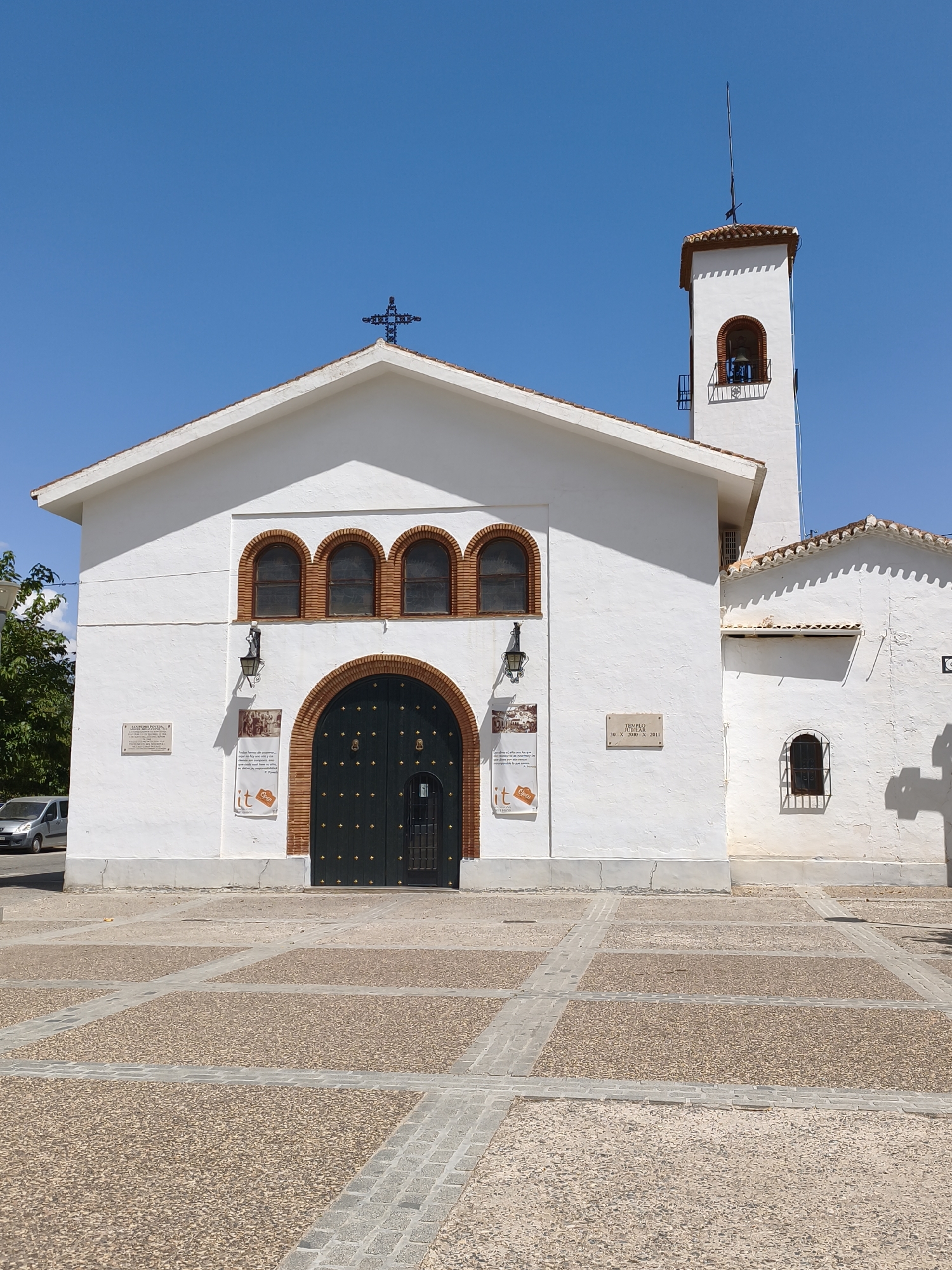 The image shows the exterior of a white church with a tiled roof under a clear blue sky. The church has a prominent arched doorway with dark green doors adorned with gold studs. Above the door are three arched windows with terracotta trim. To the right of the main building, a white bell tower with a tiled roof rises, featuring an arched window with a bell inside. A dark cross sits atop the bell tower. The church facade also has several plaques and posters. In front of the church is a paved courtyard with a pattern of light brown gravel squares bordered by grey rectangular stones. To the left of the church, a silver car is parked on a street. The scene is located in the city of Guadix, Spain.