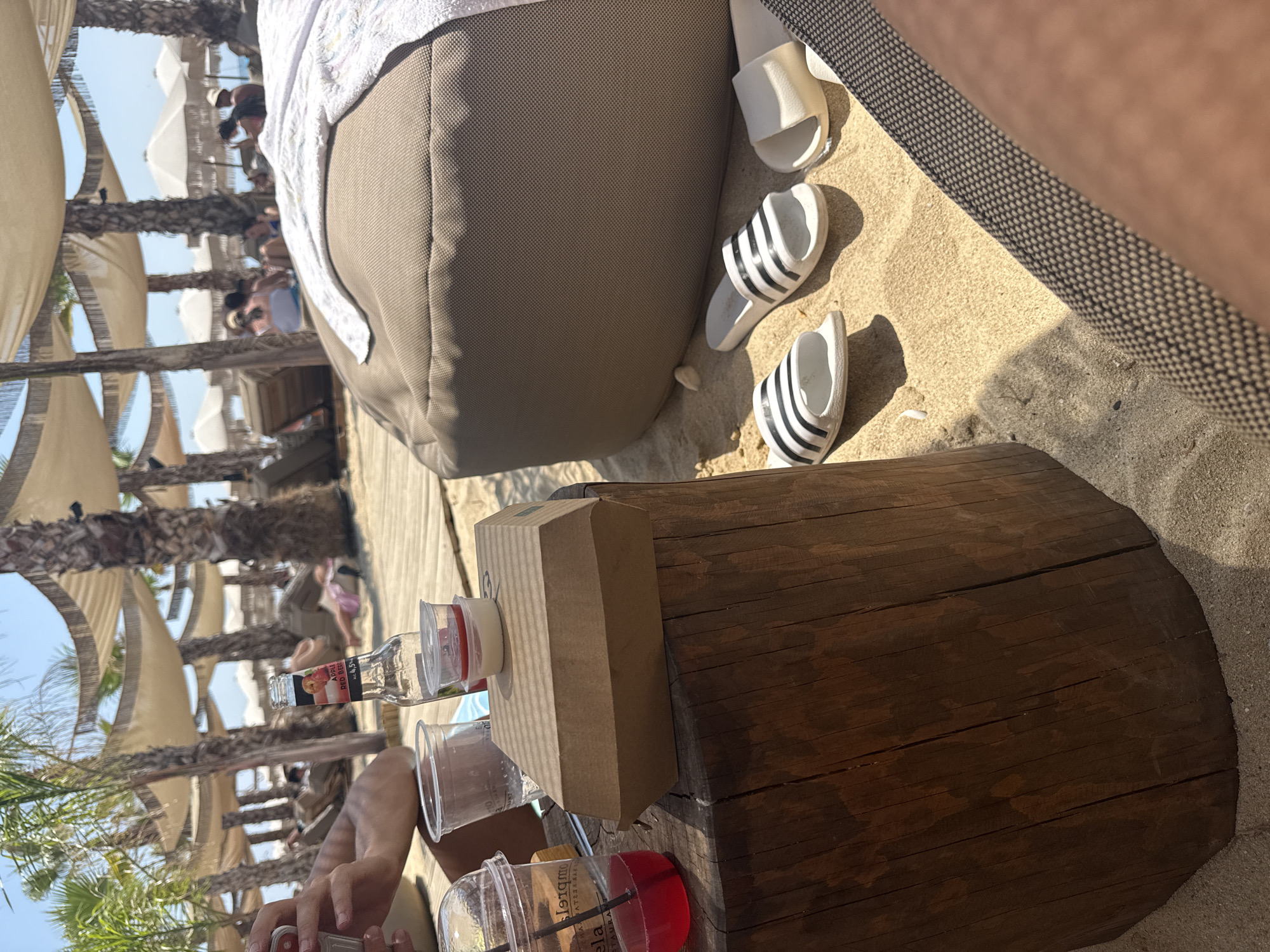 The image depicts a beachside scene at Ofrynio Beach, Greece. In the foreground, a wooden table holds several items: a plastic cup with a red beverage and a straw, a brown cardboard food container, a glass bottle of "Apple Red Berry" juice, and a small clear container with red sauce and a white dip. To the right of the table, on the sandy ground, are three pairs of flip-flops, two of which are white with black stripes, and one is solid white. A large, taupe beanbag chair with a white towel draped over it occupies the mid-ground. In the background, several palm trees with thatched canopies provide shade, with lounge chairs and umbrellas visible further back, suggesting a relaxed resort atmosphere. People are sparsely scattered throughout the background, appearing to be lounging or walking. The overall environment is sunny and appears to be a popular tourist destination.