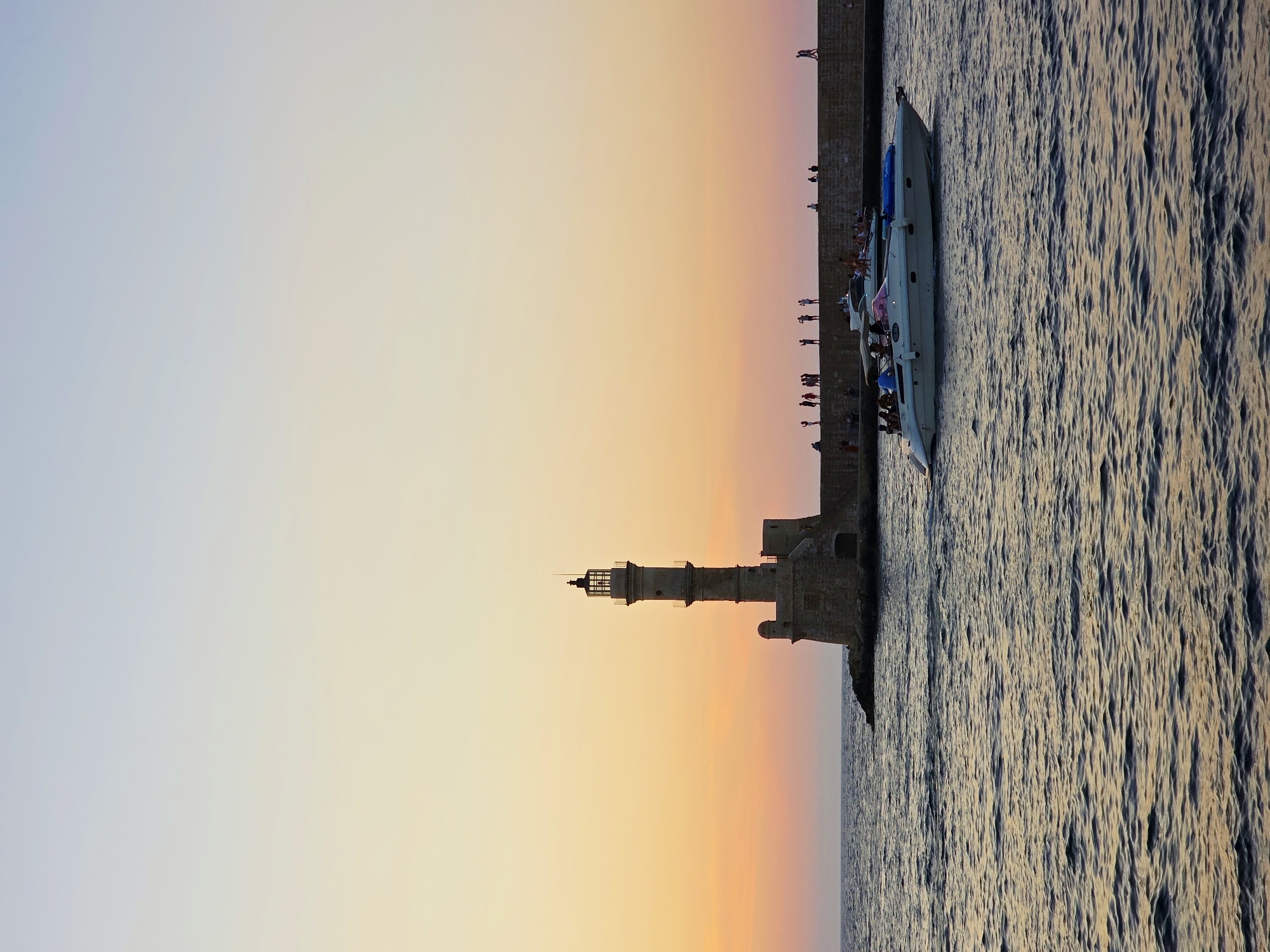The image depicts the Venetian Harbor in Chania, Greece, during sunset. A stone lighthouse, a prominent landmark, stands on a rocky outcrop to the left, with a stone breakwater extending to the right. Silhouetted figures of people can be seen walking and standing along the breakwater. In the foreground, a white motorboat with several people on board cruises on the calm, rippling sea. The sky exhibits a soft gradient of pale yellow and pink hues, characteristic of a sunset. The water reflects the muted colors of the sky and the surrounding structures.