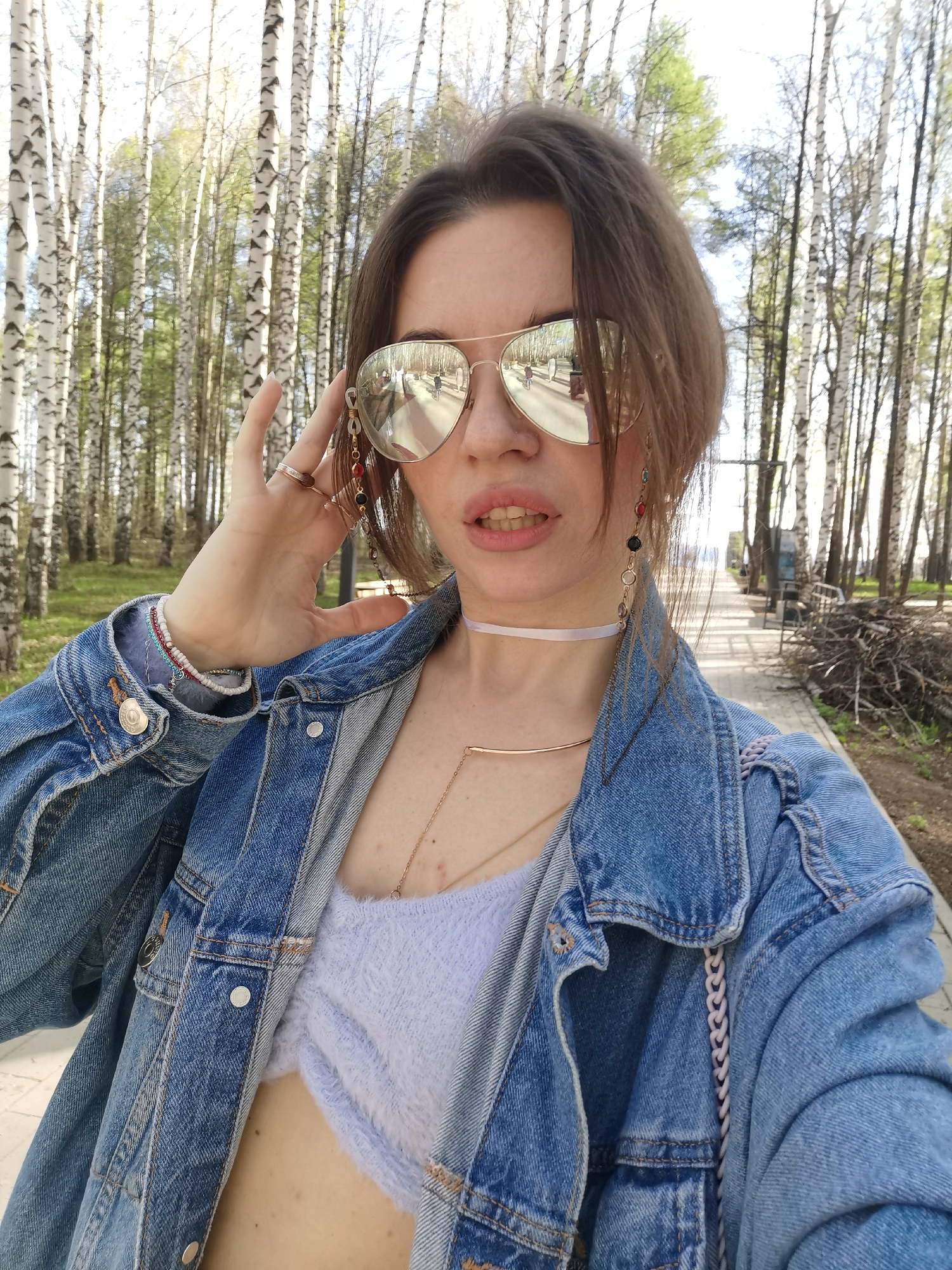 A fair-skinned woman with light brown hair and wearing reflective aviator sunglasses is the central figure in this medium shot. She is wearing a blue denim jacket over a light purple fuzzy sweater, which is partially unbuttoned to reveal some of her abdomen. Multiple pieces of jewelry adorn her: a ring on her finger, several bracelets on her left wrist, a choker around her neck, a necklace, and long earrings that are attached to her sunglasses. She is touching her sunglasses with her left hand, and looking directly at the camera.
The background features a park with many birch trees lining a paved path. In the distance, there are some benches and a small structure. To the right of the path is a pile of branches. The scene appears to be taken in a city park, under bright daylight.