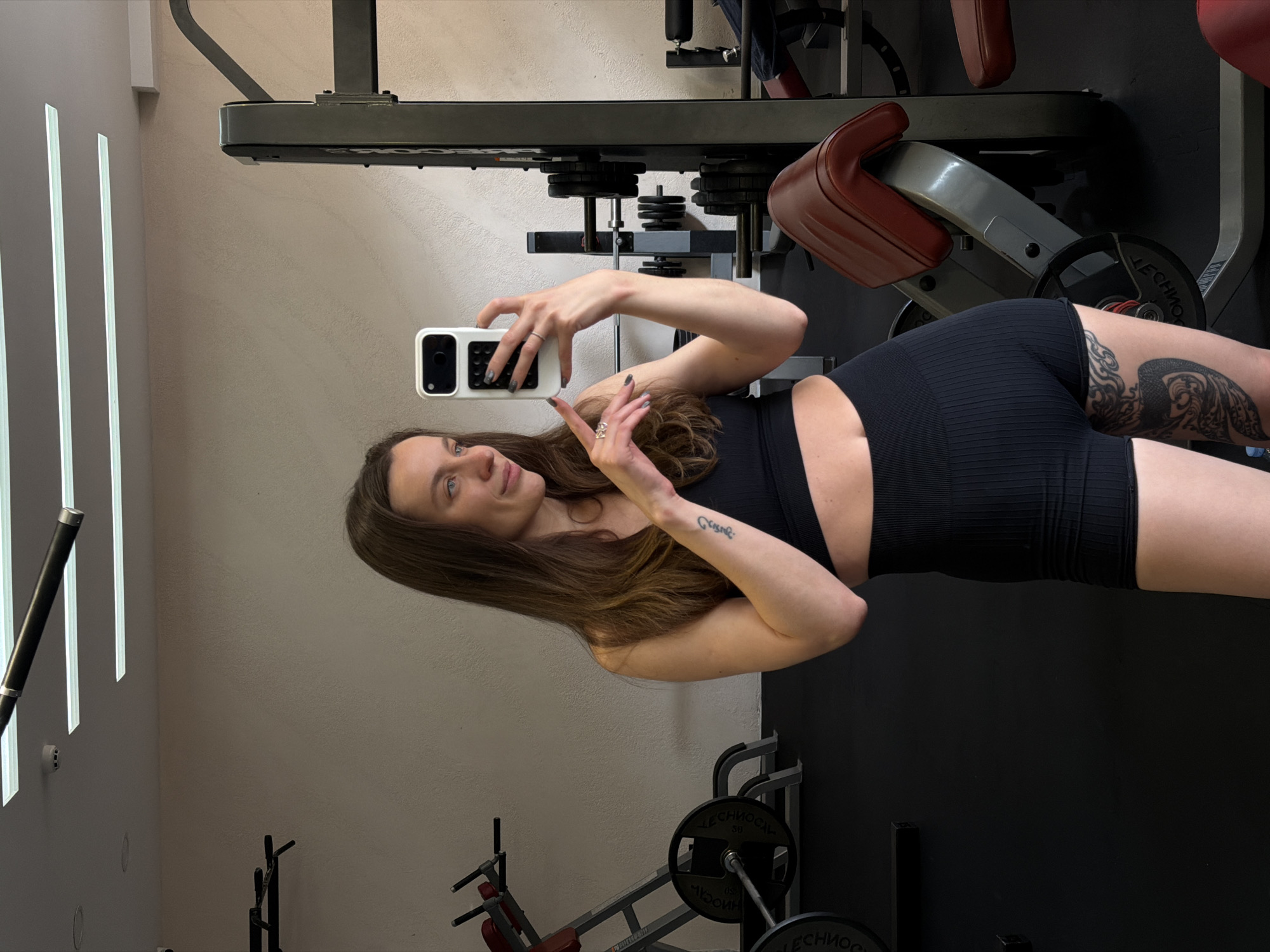 The image shows a woman in a gym. She's wearing a black sports bra and black shorts, holding a smartphone in her hand as if taking a selfie. She has brown hair and tattoos are visible on her arm and thigh. The background includes gym equipment such as weight machines and free weights. The gym has a modern look, with white walls and black accents. There is no information to indicate the location is in Budva, Montenegro.