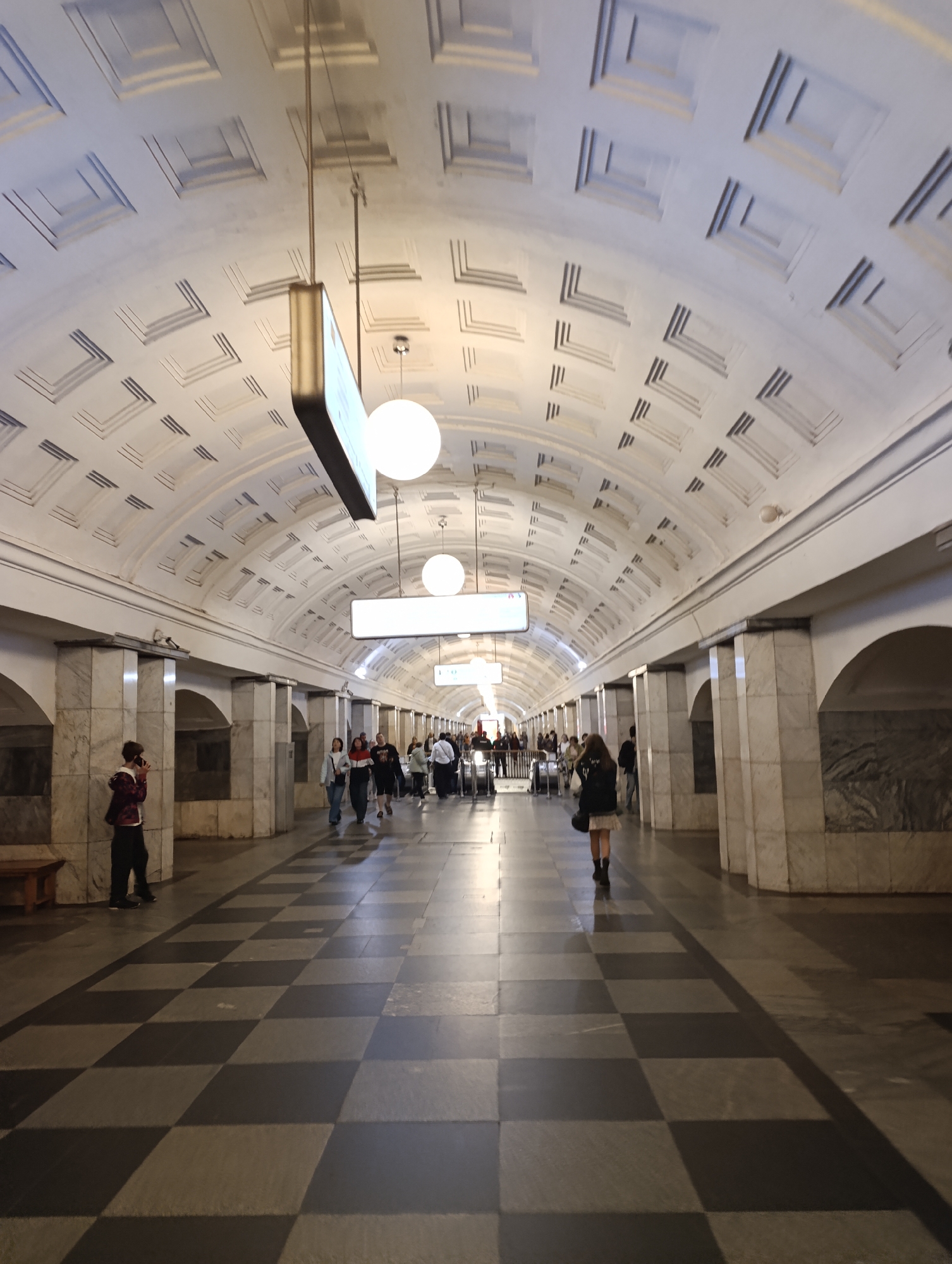 This image captures the interior of a spacious subway station platform or concourse. The scene is dominated by a grand, barrel-vaulted ceiling adorned with intricate, repetitive patterns of recessed, geometric shapes, giving it a coffered appearance. Large, circular, luminous globe lights and elongated, rectangular illuminated signs hang from the ceiling, providing ample artificial light.
The station's architecture features sturdy columns and wall sections clad in light-colored marble or stone, with darker, possibly granite or polished stone, panels interspersed along the lower walls and within arched recesses. A prominent feature is the checkered floor, composed of alternating black and white squares, which stretches into the distance, creating a sense of depth and perspective.
Several people are visible throughout the station, engaged in various activities typical of a public transport hub. Some individuals are walking along the platform, while others appear to be waiting or conversing. On the left side, a person in a plaid shirt is seen standing near a wooden bench, holding a phone to their ear. Further down the platform, a cluster of people approaches what appears to be an escalator system in the background. The general atmosphere suggests a moderately busy period.
The lighting is consistent and even, typical of an indoor, underground environment. There is no natural light, and the time of day is not discernible. The illuminated signs hanging from the ceiling are too distant to reveal any legible text. The overall impression is one of architectural grandeur and functional public space.
