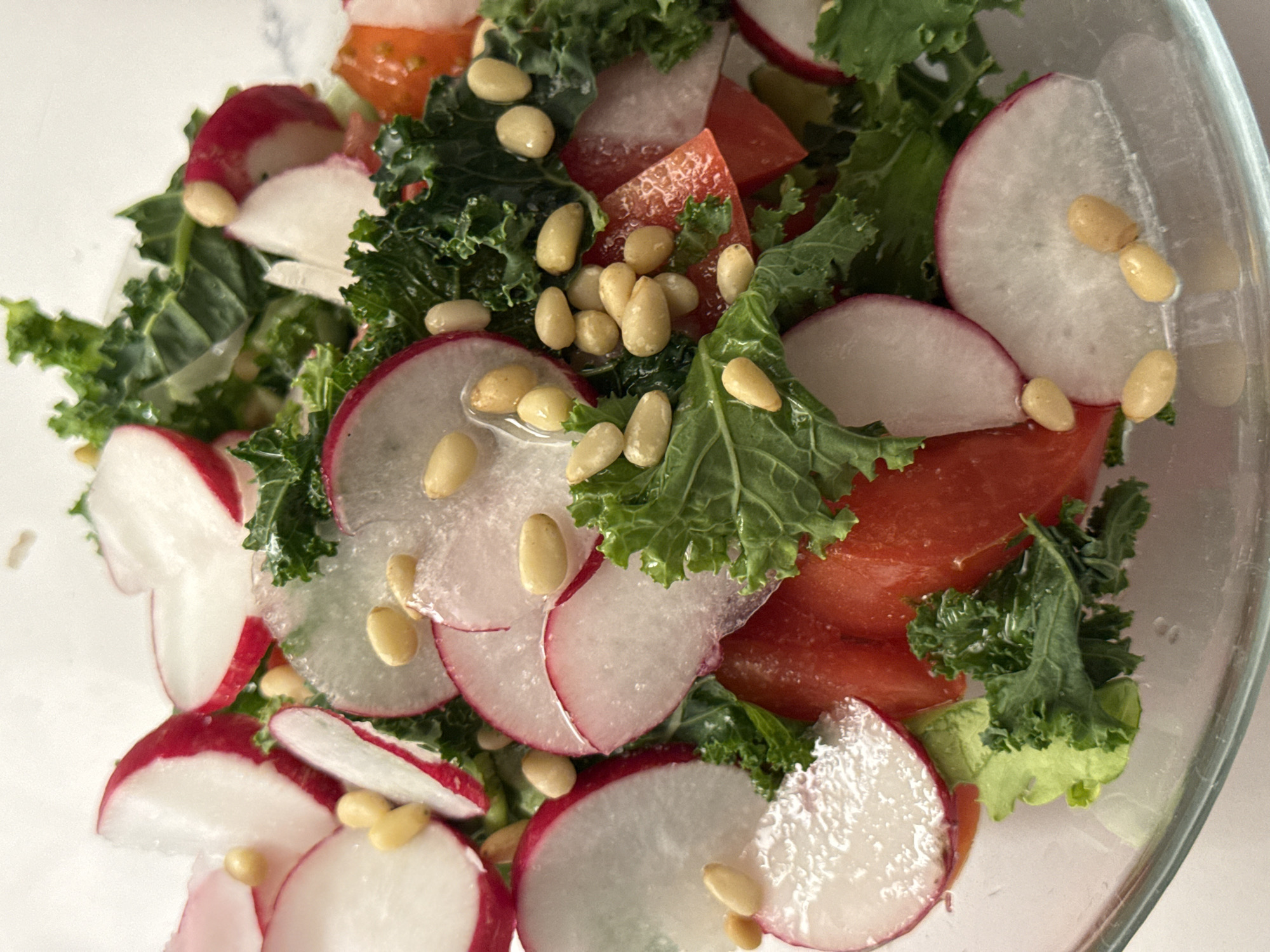 A close-up shot shows a salad in a glass bowl. The salad includes sliced radishes with red skins, pieces of tomatoes, kale, and pine nuts. The environment appears to be a brightly lit indoor space. No people are visible. The location in the city of Moscow, Russia is not discernible from the given image.