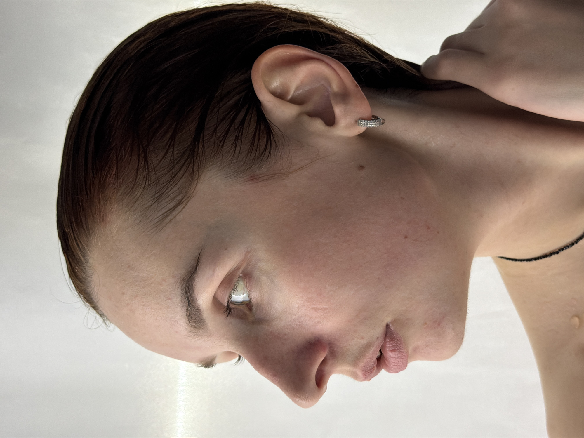 A close-up, tilted headshot of a fair-skinned person with dark, wet hair. They are wearing a small, hoop earring in their left ear and a delicate, black beaded necklace. The background is a soft white, possibly a solid surface or wall. The person's right hand is raised near their neck. A single droplet of water is visible on their right shoulder. The location is not discernible from the image.