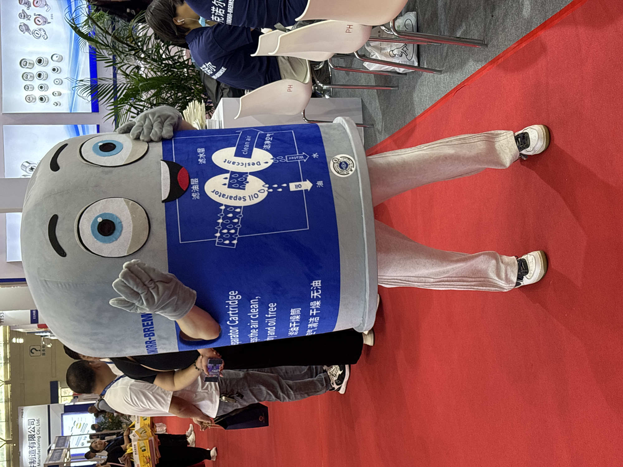 The image shows a mascot in a blue and gray costume, resembling a filter cartridge with large cartoon eyes and gray gloves. The costume is labeled "Oil Separator Cartridge" and "NORR-BREM" and has a diagram indicating oil, water, and desiccant filtration. The person wearing the costume has light-colored pants and white sneakers, and is standing on a red carpet. Several people are gathered around, some taking pictures. The environment appears to be a trade show or exhibition in the city of Liangjiang, China, with other displays and exhibitors visible in the background, including booth signage in both English and Chinese. Chairs labeled "PH" are in the background.