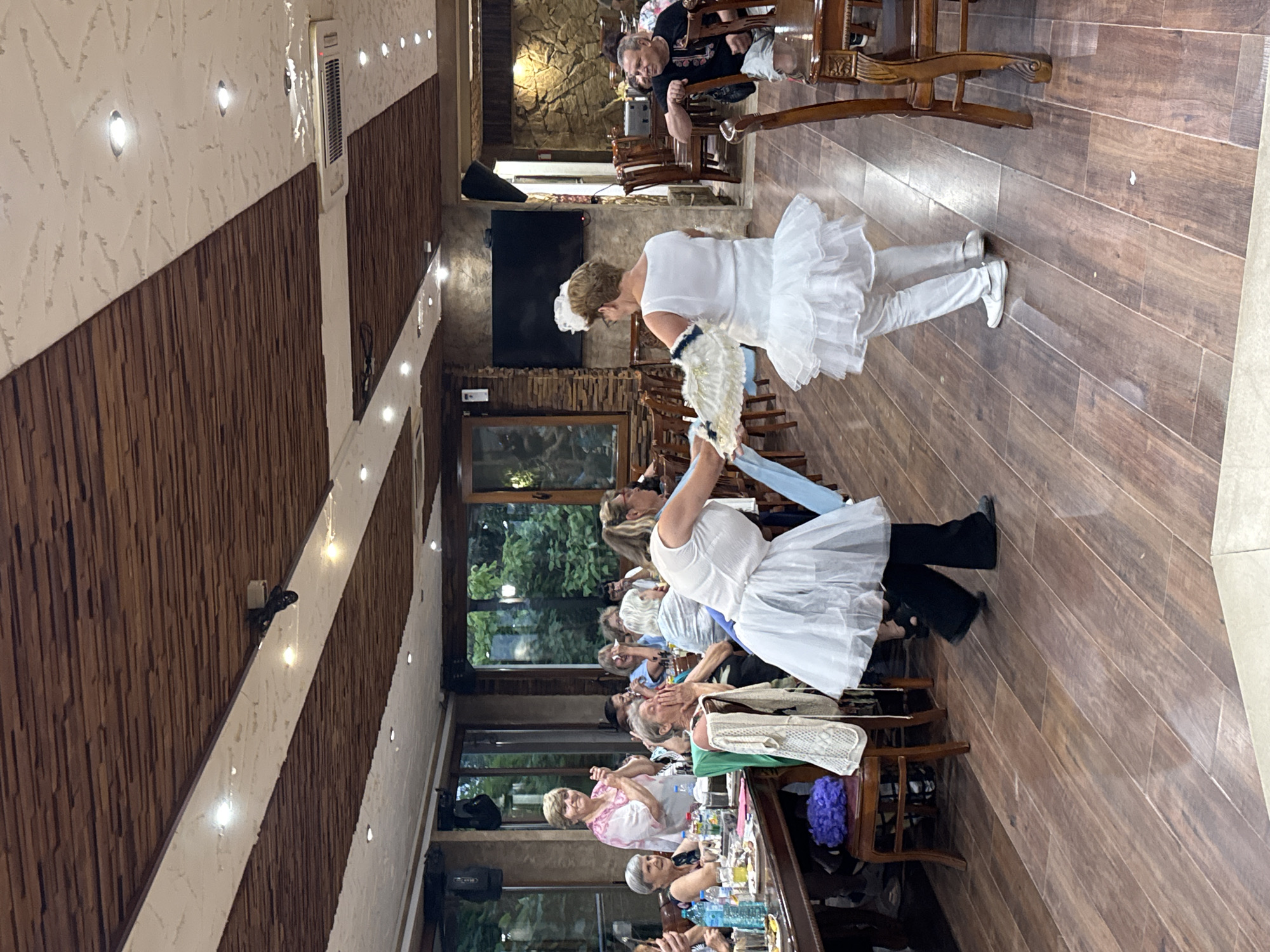 The image depicts an indoor scene, likely a restaurant or event hall, with a wooden ceiling and floor. The setting appears to be Sredets, Bulgaria, as indicated by contextual information.
In the foreground, two individuals are engaged in a performance or dance. One person, wearing a white layered skirt and white leggings, is positioned with their back to the camera, holding a fan. Another person, also in a white layered skirt and black pants, is facing the first individual, holding a blue sash or fabric. Their attire suggests a coordinated performance.
Surrounding them are several tables with diners, many of whom are looking towards the performers. There are bottles of drinks and plates on the tables. The diners are a mix of ages and genders, some appearing to be clapping or reacting to the performance.
In the background, there are more tables and chairs, along with windows that offer a view of greenery outside. The walls are decorated with stone and wooden elements, and a television screen is visible on one wall. Lighting fixtures are integrated into the ceiling. The overall atmosphere suggests a social gathering with entertainment.