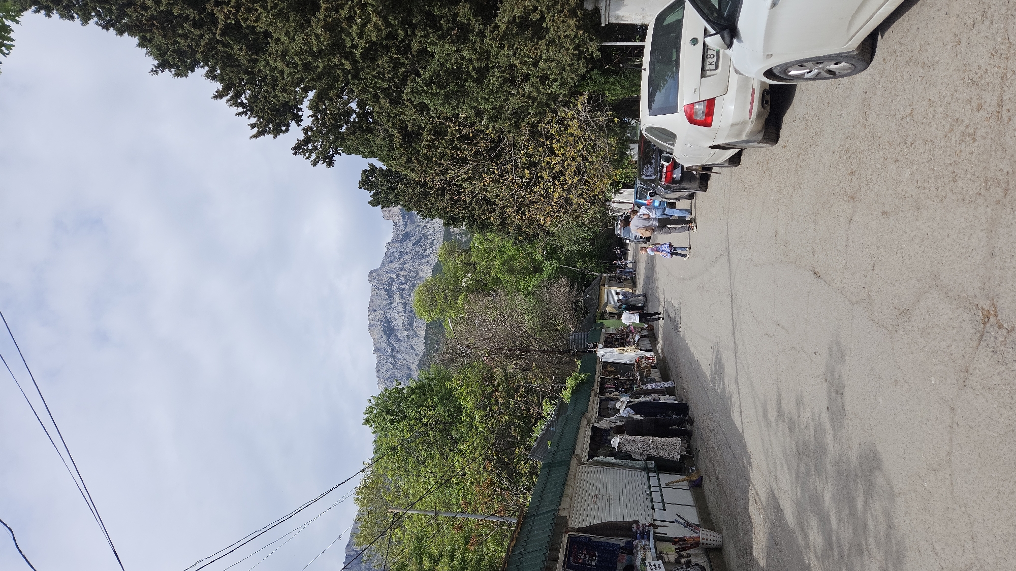 A street scene in the city of Unknown, Unknown, shows a road lined with stalls selling goods, with people walking and browsing. Cars are parked along the side of the road. In the background, a mountain looms over the scene, and trees frame the view. Power lines stretch across the sky. The road surface is marked with shadows and imperfections.