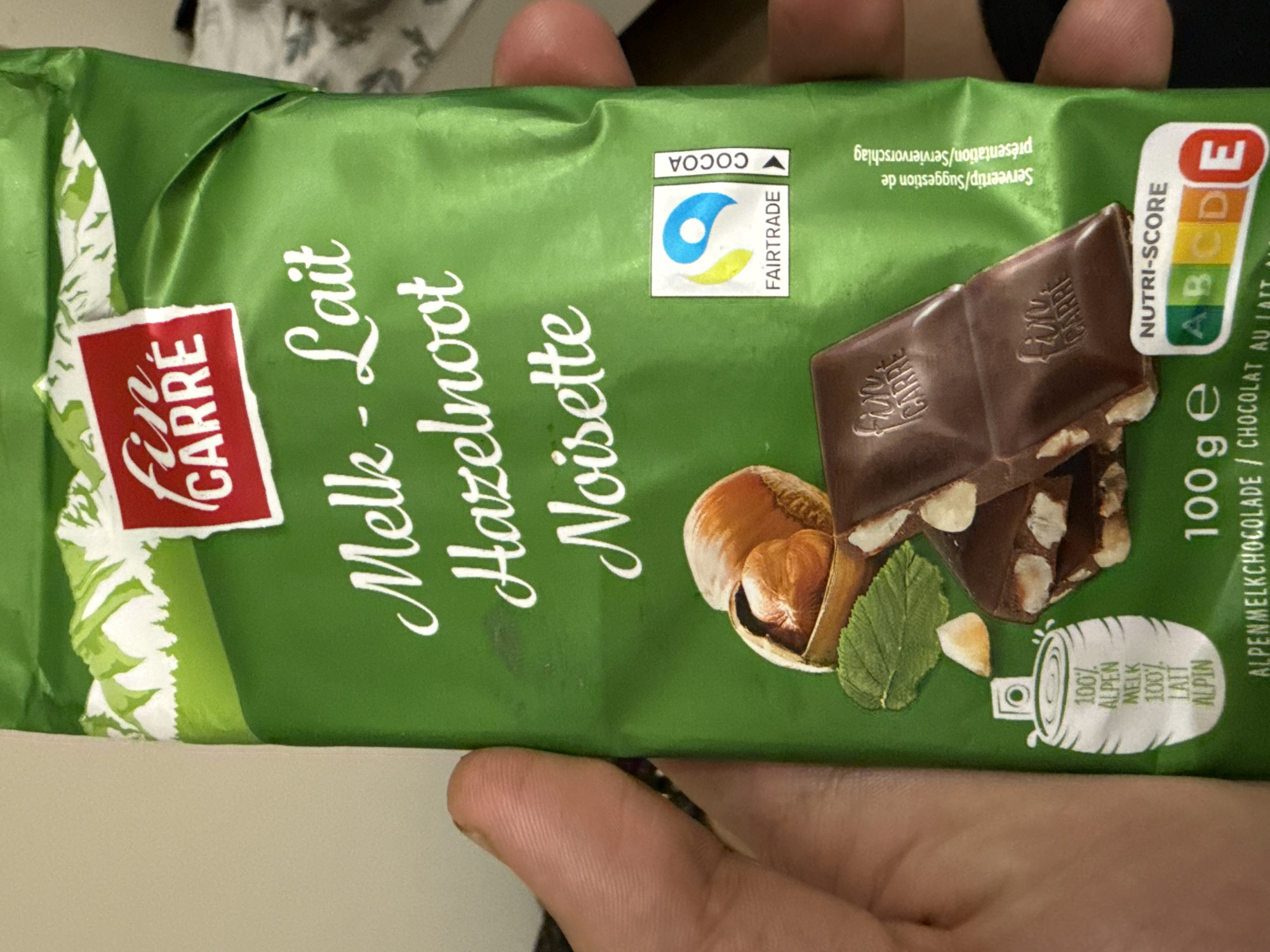 A person's hands are holding a green Fin Carré brand milk chocolate bar with hazelnuts. The hands are visible at the top and bottom of the frame, holding the package horizontally, suggesting an indoor setting in Dobrogled, Bulgaria. The chocolate bar wrapper features various texts and logos.
Prominently displayed on the wrapper is the brand name "Fin CARRÉ" on a red label, along with "Melk - Lait Hazelnoot Noisette," indicating milk chocolate with hazelnuts in Dutch and French. An illustration shows several pieces of chocolate with visible hazelnut chunks, alongside whole hazelnuts and a mint leaf. The chocolate pieces themselves have "Fin CARRÉ" embossed on them.
A "FAIRTRADE" logo is visible, with "COCOA" written above it, signifying fair trade certified cocoa. A "NUTRI-SCORE" label is present, displaying an 'E' score, indicating its nutritional rating. Other text includes "Serveertip/Suggestion de présentation/Serviervorschlag" (serving suggestion) and "100 g e" (weight). A graphic with "100% ALPEN MELK 100% LAIT ALPIN" suggests the use of Alpine milk. The background is blurred, offering no specific details about the immediate surroundings beyond it being an indoor environment.