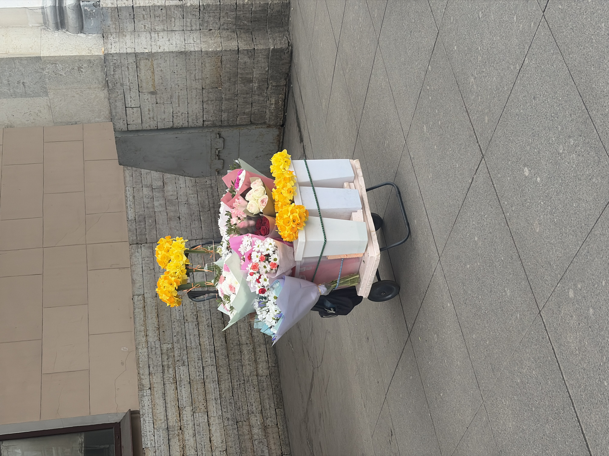 An outdoor urban scene in Saint Petersburg, Russia, featuring a mobile flower stand. The main subject is a two-wheeled, hand-pulled metal cart with a pale pink wooden platform, displaying numerous bouquets of fresh flowers.
**Subjects and Objects:**
* **Flower Cart:** The cart has a dark metal frame, two black wheels, and a rectangular, light pink wooden platform.
* **Flowers:** The platform holds three white rectangular plastic buckets containing bundles of bright yellow daffodils. Additionally, various bouquets are arranged on and around the cart, wrapped in pastel-colored paper (pink, light blue, purple, white). These bouquets consist of mixed flowers, including white and red daisies, pink and white roses, and other mixed floral arrangements. A black soft-sided bag is suspended from the cart's side handle.
**Environment and Location:**
* **Ground:** The cart rests on a paved sidewalk composed of large, uniform grey square tiles.
* **Background (Left):** The lower section of a building wall features several courses of light-colored, roughly cut stone blocks laid horizontally. Above this stone base, the wall transitions to a smooth, light beige facade with a visible dark-framed window, angled downwards from the top left corner.
* **Background (Right):** A large, light-colored classical column base, exhibiting decorative molding and visible staining/wear, rises from the stone-block wall. Adjacent to the column base, another section of the wall is constructed from rough-hewn stone blocks. A dark grey, slightly slanted, rectangular structure is visible in the mid-ground, positioned between the stone wall sections.
* **People/Actions:** No individuals are present in the image. The scene is static.
**Location Detail:**
The architectural elements, including the heavy stone block bases, the classical column, and the grand scale of the building facades, are characteristic of the historic center of Saint Petersburg, Russia. Specifically, this environment strongly aligns with structures found along **Nevsky Prospekt** or in its immediate vicinity, such as the area around Gostiny Dvor, known for its neoclassical architecture and common presence of street vendors.