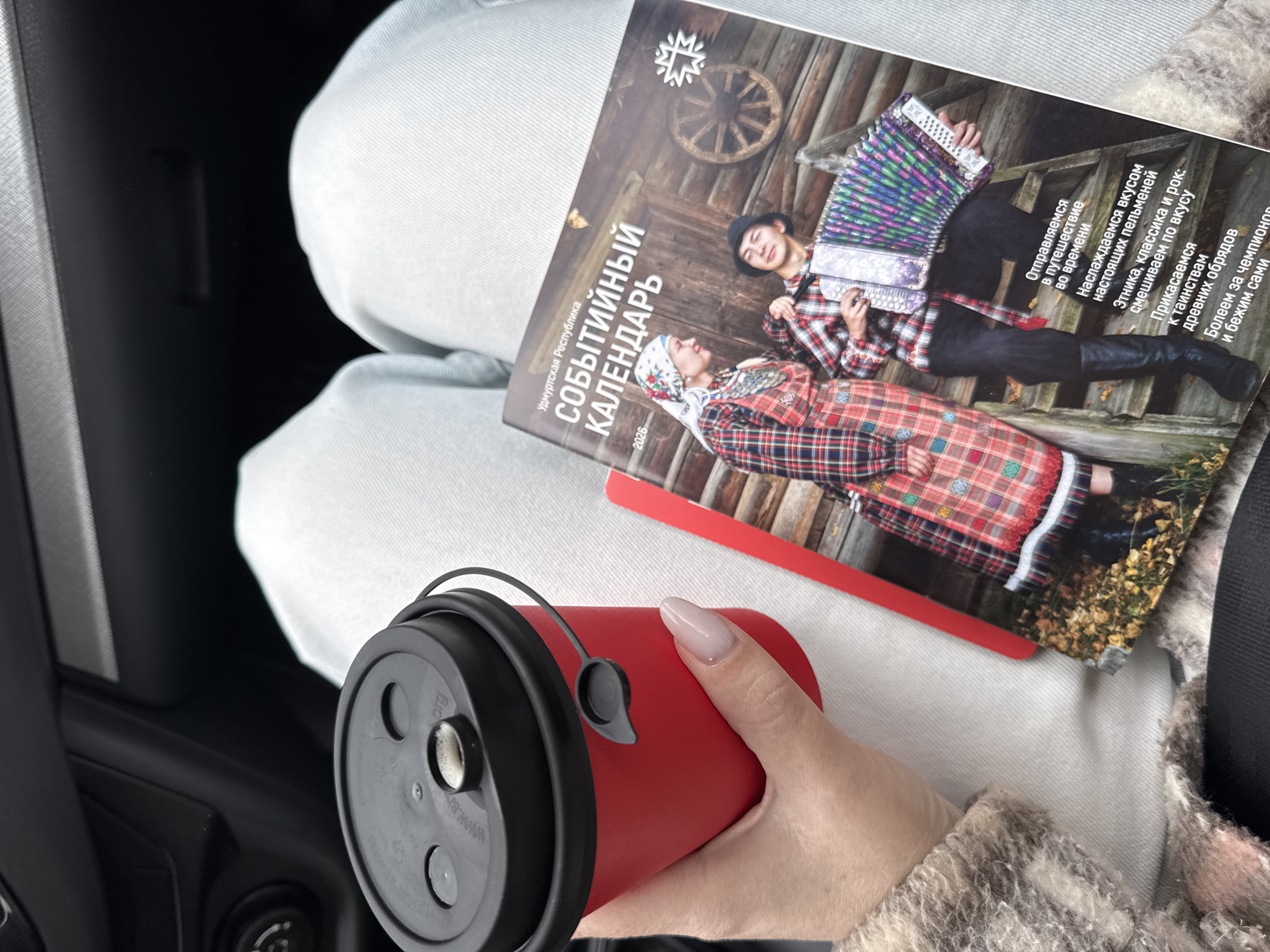 Here is a concise description of the image:
The image shows a person sitting with a red travel mug in their hand. Resting on their lap is a calendar for the Republic of Udmurtia, Russia, specifically the year 2026. The calendar features two people in traditional dress. Votkinsk is not mentioned.