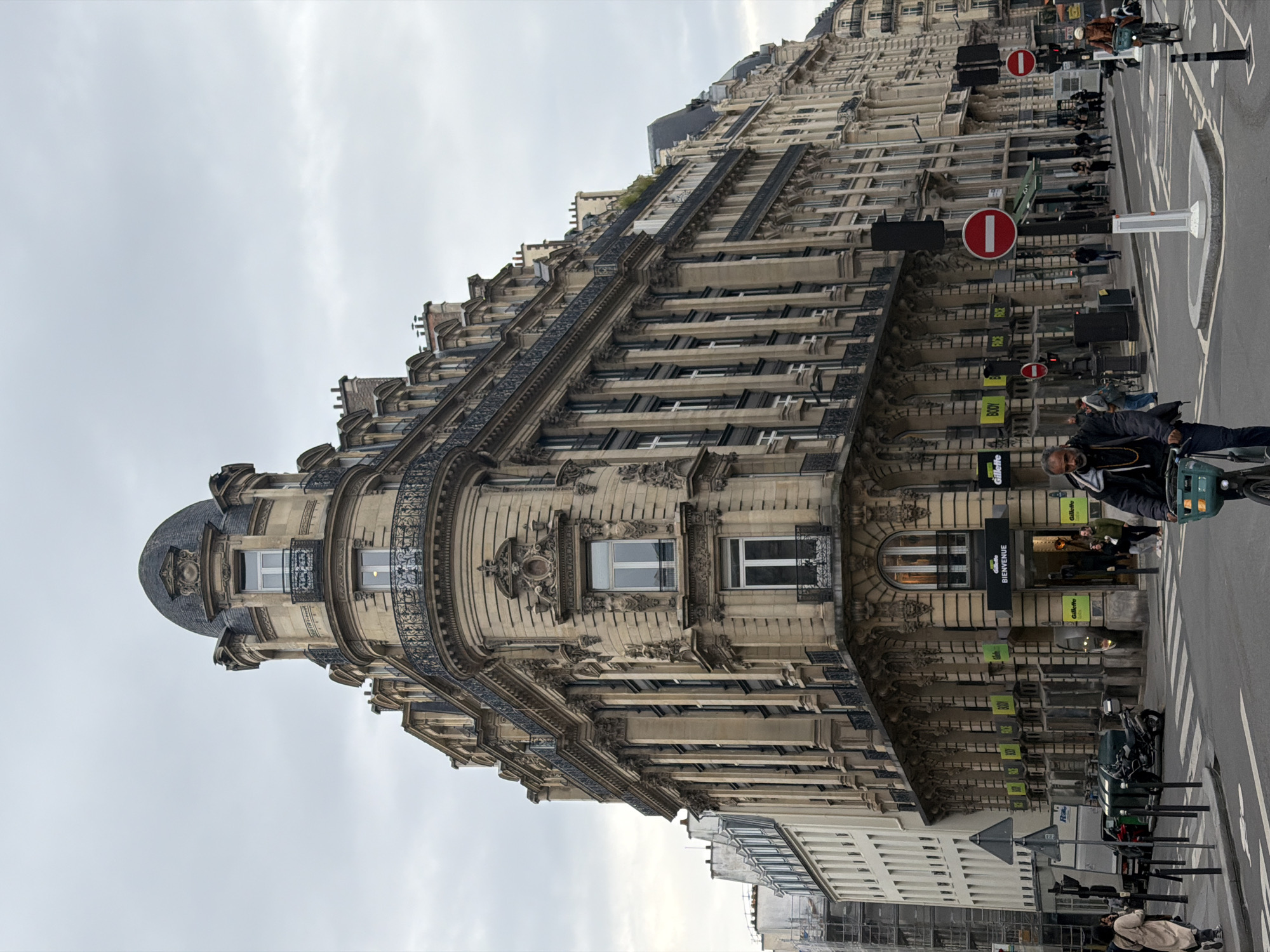 The image displays a street scene at the intersection of Boulevard des Capucines and Rue des Capucines in Paris, France, under an overcast sky. A prominent five-story Haussmannian building, constructed of light-colored stone, dominates the foreground, featuring a distinctive domed corner turret and decorative wrought-iron balconies. The building's ground floor houses a "Gillette" retail store, indicated by black signage bearing the "Gillette" logo and the word "BIENVENUE" (French for "Welcome") above the main entrance. Additional "Gillette" branding and signs for "BODY," "FACE," and "HAIR" are visible along the storefront façade.
To the right, a continuous row of similar ornate Haussmannian buildings extends down the street. To the left, a plainer, lighter-colored building with visible scaffolding on its upper levels is partially seen. The street intersection is paved with asphalt, marked by white lane lines and pedestrian zebra crossings. A concrete island with a black traffic signal pole and a red circular "no entry" sign is centrally located in the foreground. Another "no entry" sign and traffic lights are visible further along the street to the right.
Multiple individuals are present. In the immediate foreground, a man with a beard, wearing a dark jacket over a hoodie, rides a bicycle equipped with a green front basket, moving towards the left of the frame. Other pedestrians are distributed across the sidewalks and crossing the street; one woman in a light coat walks away on the left sidewalk. A second cyclist is visible further down the street on the right. A dark-colored scooter is parked on the left side of the street, near a white truck. The scene is illuminated by diffused natural light, consistent with an overcast daytime setting.