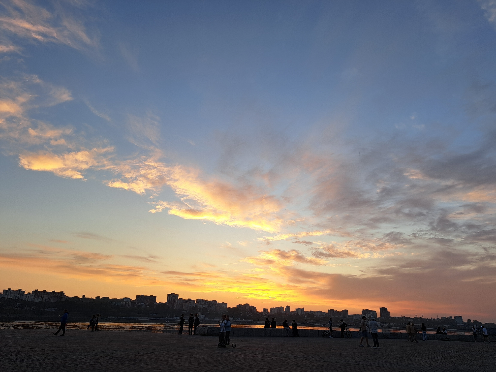 A wide, horizontal, eye-level shot captures a sunset over the Angara River in Irkutsk, Russia. The sky is a gradient of soft blues and oranges, with streaks of clouds catching the warm light. In the foreground, a paved walkway is populated by several individuals and couples, some standing and some walking. Among them, a person on a scooter stands out. Beyond the walkway, the dark silhouette of the Angara River reflects the sky's colors. Across the river, the cityscape of Irkutsk is visible, characterized by a skyline of residential buildings and apartment complexes. The overall scene is serene, with a sense of quiet observation of the natural phenomenon.