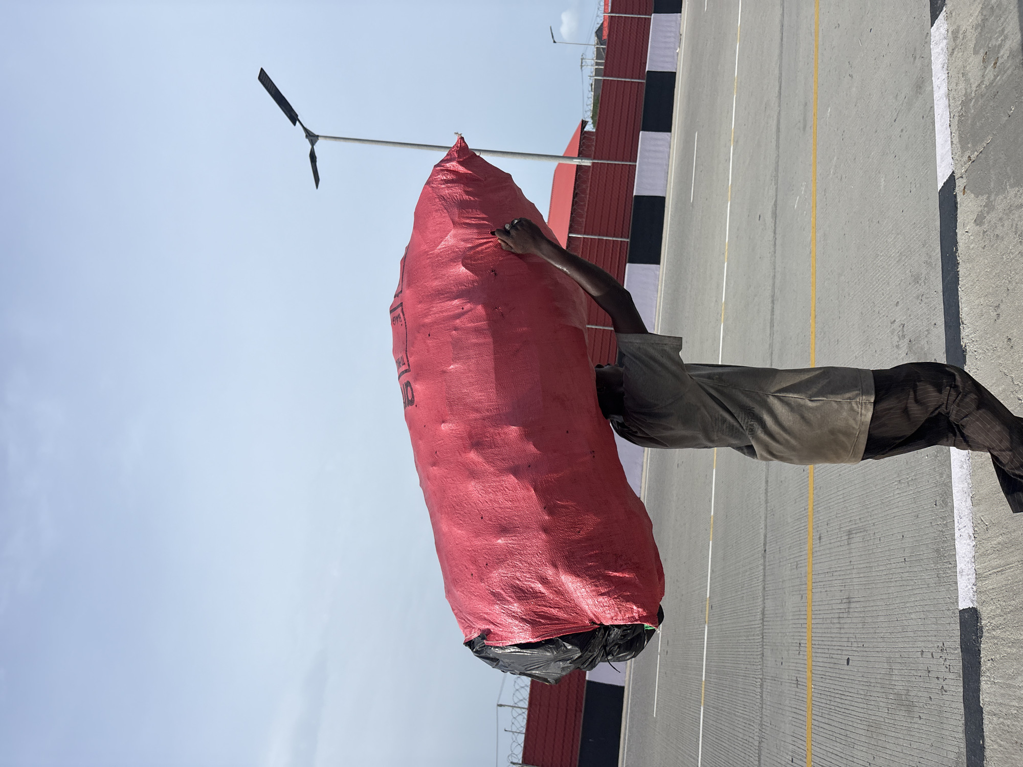 The image shows a man carrying a large red sack on his shoulder along a concrete road in Alaguntan, Nigeria. The road has yellow and white lines dividing the lanes, and a wall with black and white squares runs alongside it. A streetlight is visible in the background. The man appears to be walking with the sack resting on his shoulder, possibly transporting goods or materials.