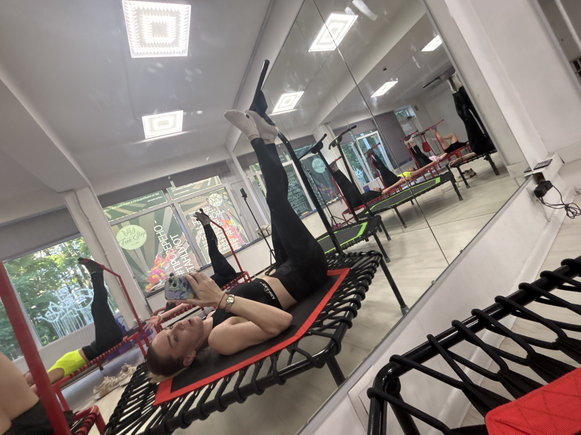The image shows an indoor fitness studio with multiple mini-trampolines. In the foreground, a woman is lying on her back on a black mini-trampoline with a red edge. She is wearing a black sports bra and black leggings, and has white socks on her feet. She is holding a smartphone in her hands, looking down at it. Her head is turned towards the camera. The studio has a mirrored wall, reflecting other people on mini-trampolines in various exercises, as well as fluorescent ceiling lights and windows with trees visible outside. The overall impression is a class in progress. The text on the windows and signs suggests the location is in Sinyavino, Russia.