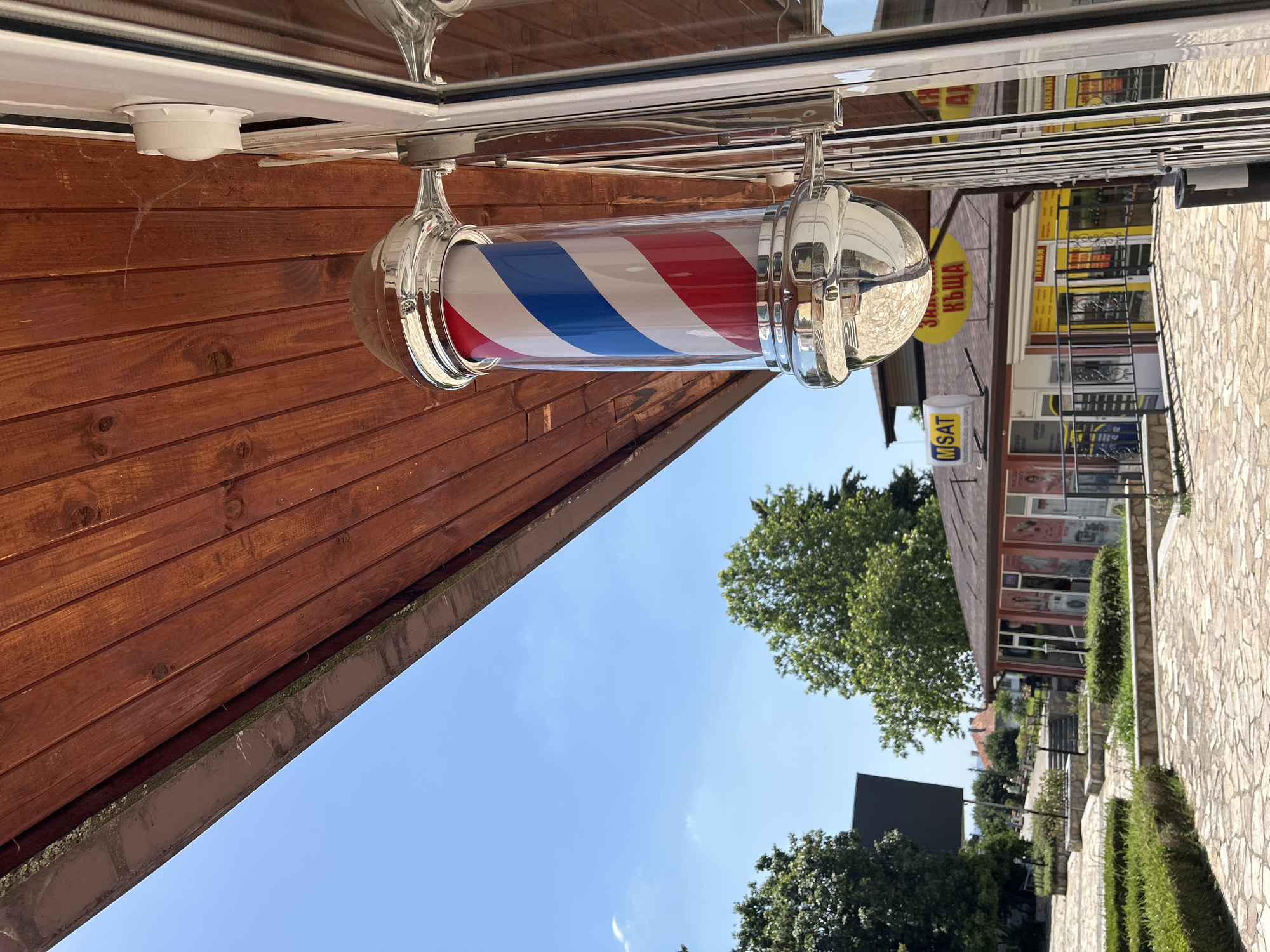 This media file displays an outdoor scene featuring a traditional barber pole with red, white, and blue stripes, suspended from a white awning. The barber pole is a cylindrical glass tube with chrome accents. The background reveals a building with shops, some with yellow awnings and signage. One sign is partially visible with Cyrillic text. A ladder is leaning against one of the buildings. To the right, a stone-paved walkway runs alongside manicured green hedges. Trees and a clear blue sky with a few wispy clouds are visible above the buildings and walkway. The image appears to be taken on a sunny day. Visible text includes "MSAT". The orientation of the image is rotated.