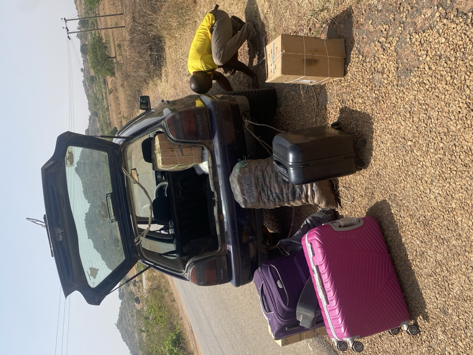 This image, taken in Zankan, Nigeria, depicts a dark-colored hatchback car parked on a gravel shoulder adjacent to an asphalt road under a clear, bright sky. The car's trunk is open, revealing a large, rope-secured cardboard box and other contents. A single adult male, wearing a yellow short-sleeved shirt and light-colored trousers, is in a crouched position near the car's rear-right wheel, appearing to inspect or attend to something beneath the vehicle. Scattered on the gravel around the car are several items of luggage and cargo: a large purple wheeled suitcase, a large pink wheeled suitcase, a smaller black wheeled suitcase, a tall burlap sack labeled "PORKOS," and a rope-bound cardboard box. The background features sparsely vegetated hills and power lines with poles.