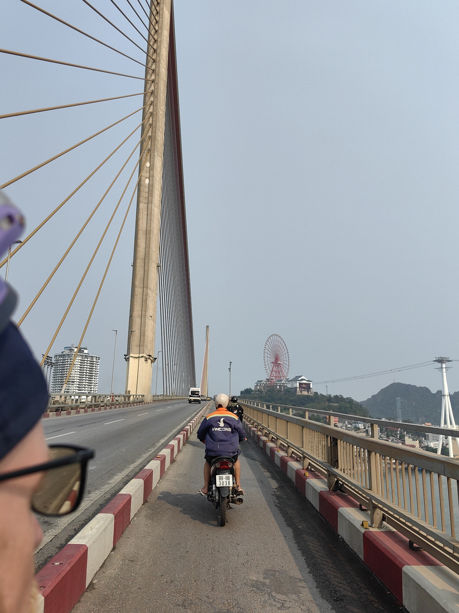 A photograph captures a bridge in Bai Chay Ward, Vietnam. A tall bridge tower is prominently featured to the left of the frame, with visible cables extending outwards. A Ferris wheel is visible in the distance. A motorcycle with two riders is traveling on the bridge. A pedestrian with sunglasses and a hat is partially visible in the foreground. A vehicle can be seen driving on the opposite side of the bridge. A body of water is visible beyond the bridge's railing, with a cable car structure spanning across. Buildings are visible in the background on the distant shore.