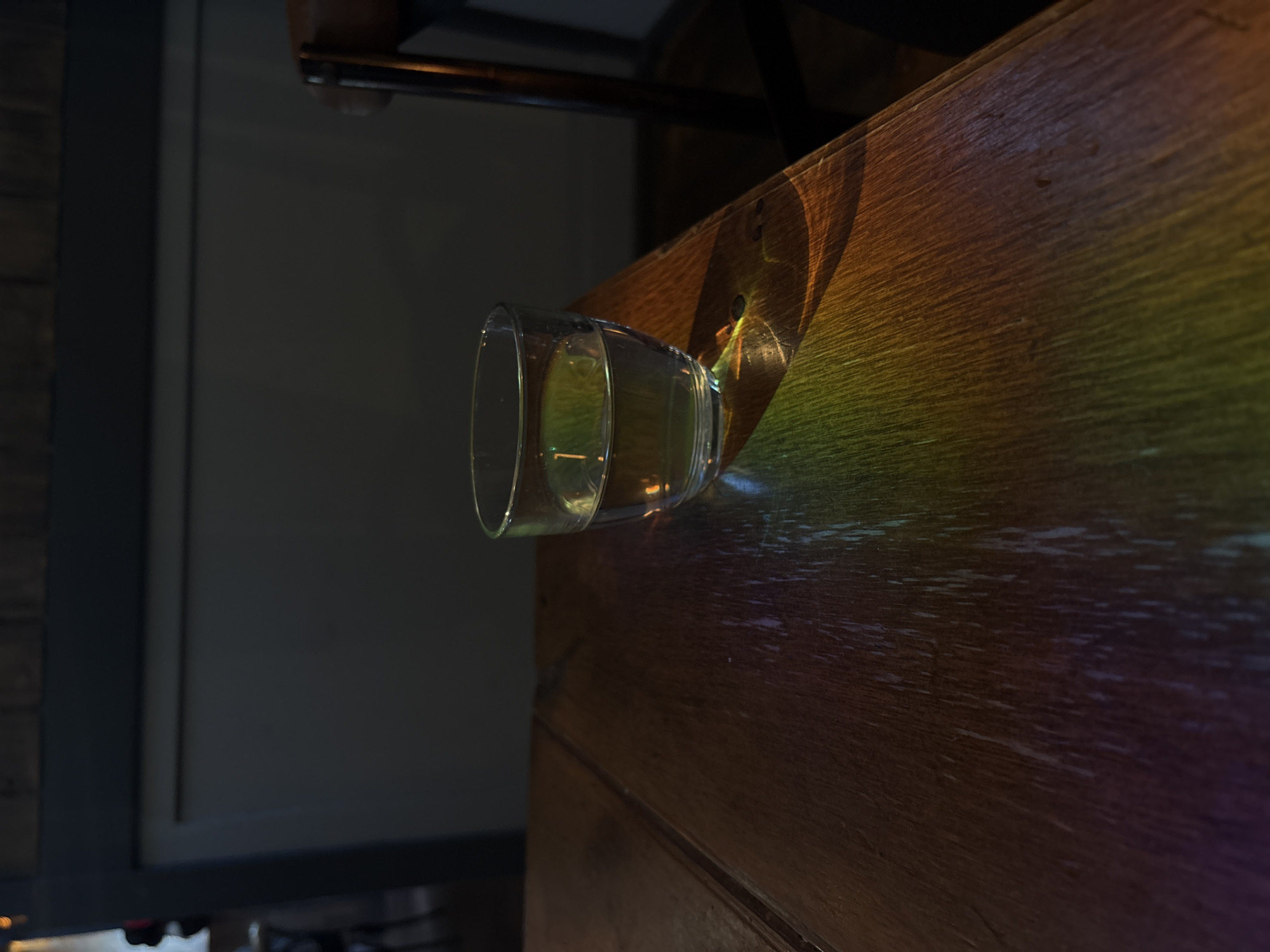 A clear glass of water sits on a wooden table, casting a rainbow of light onto the surface. The table appears to be in a dimly lit room, possibly a bar or restaurant, given the presence of a wooden chair in the background and the dark, textured wall behind it. The lighting creates a dramatic effect, highlighting the water in the glass and the grain of the wood. The scene is still and quiet, suggesting a moment of contemplation or a pause in activity. The location is specified as Ipswich, United Kingdom.