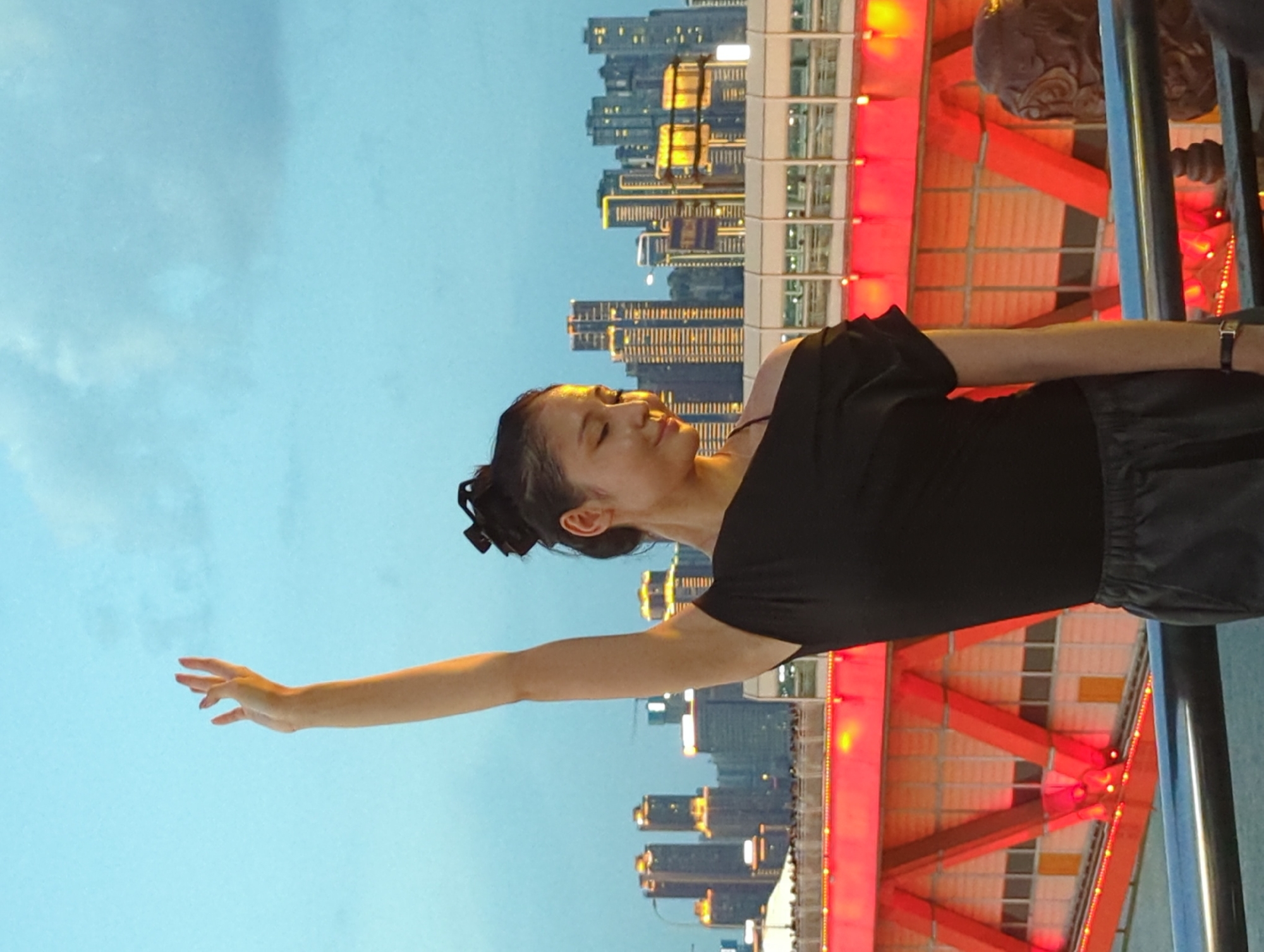 A young woman with dark hair pulled up in a bun is posing on a balcony with a city skyline and a bridge in the background. She is wearing a black off-the-shoulder top and grey shorts. Her right arm is raised in the air. The bridge, likely the Hongyadong bridge in Yuzhong District, China, is illuminated with red lights. The sky is a dusky blue with some clouds. The city skyline features several tall buildings with lights on.