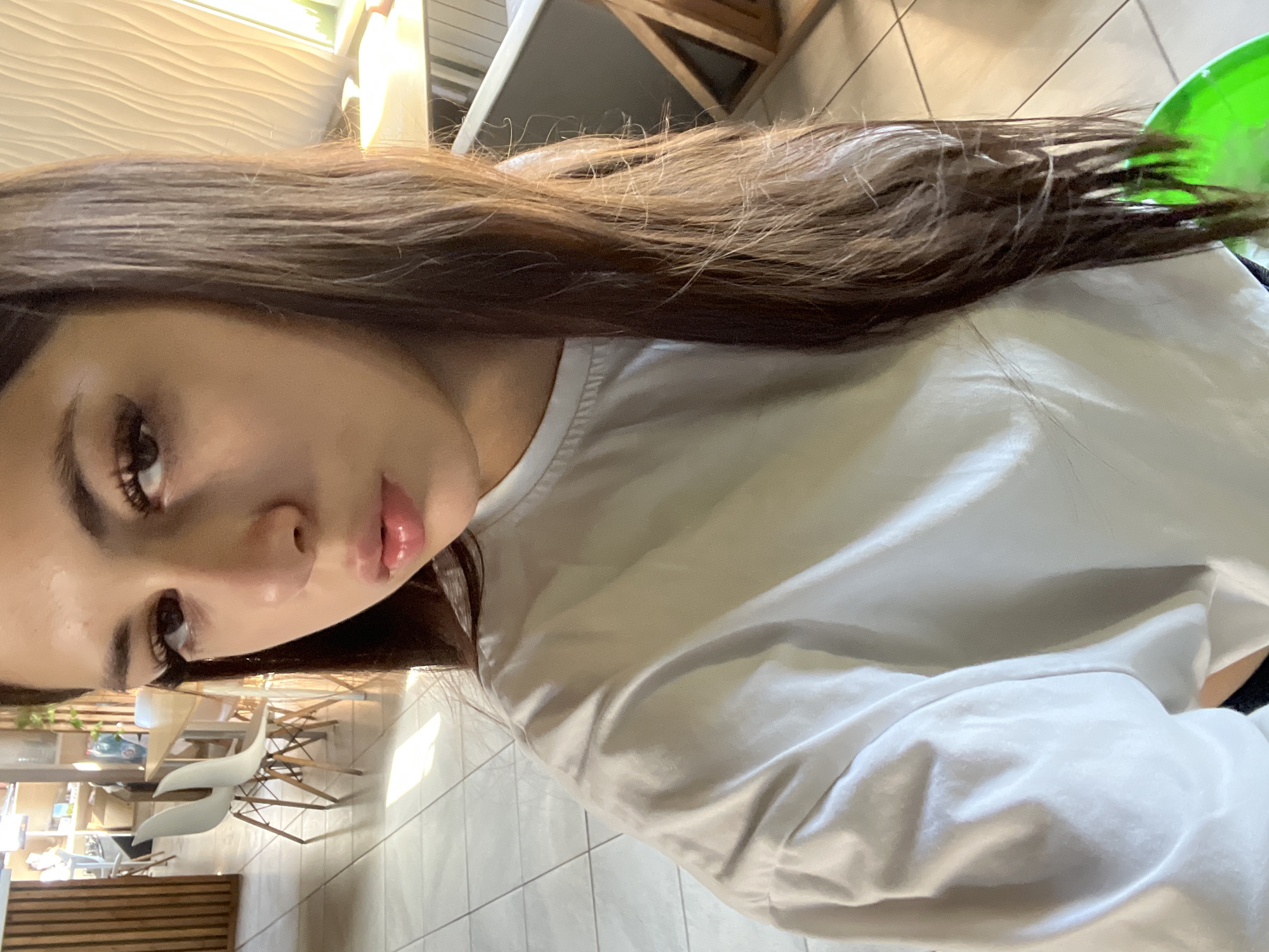 A young woman with long, dark hair is taking a selfie in what appears to be a cafe or restaurant. She is wearing a white t-shirt. Her hair is partially obscuring her face. The background shows tables and chairs, and large windows letting in natural light. The floor is tiled. A bright green object is visible at the bottom of the frame. There is no visible indication of the specific city of Krasnye Baki, Russia.