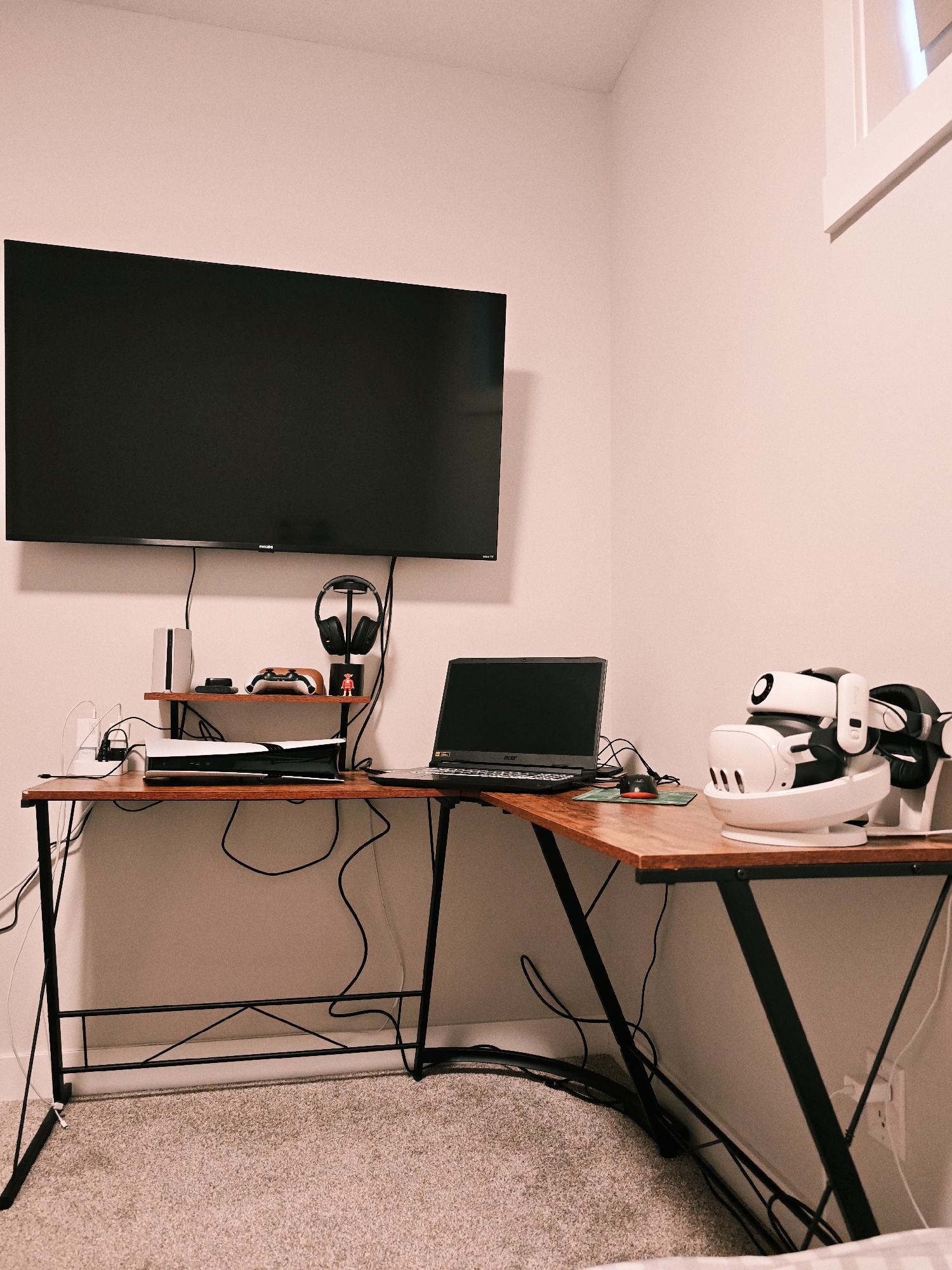 This image shows a home office setup in Calgary, Canada, featuring a corner desk with a computer, gaming console, and virtual reality equipment. A large flat-screen television is mounted on the wall above the desk. The desk itself is made of wood with black metal legs and houses a laptop, a PlayStation 5 console, and a set of headphones on a stand. On the right side of the desk, a white and black virtual reality headset is displayed on a charging dock. The room has light pink walls and a beige carpet.