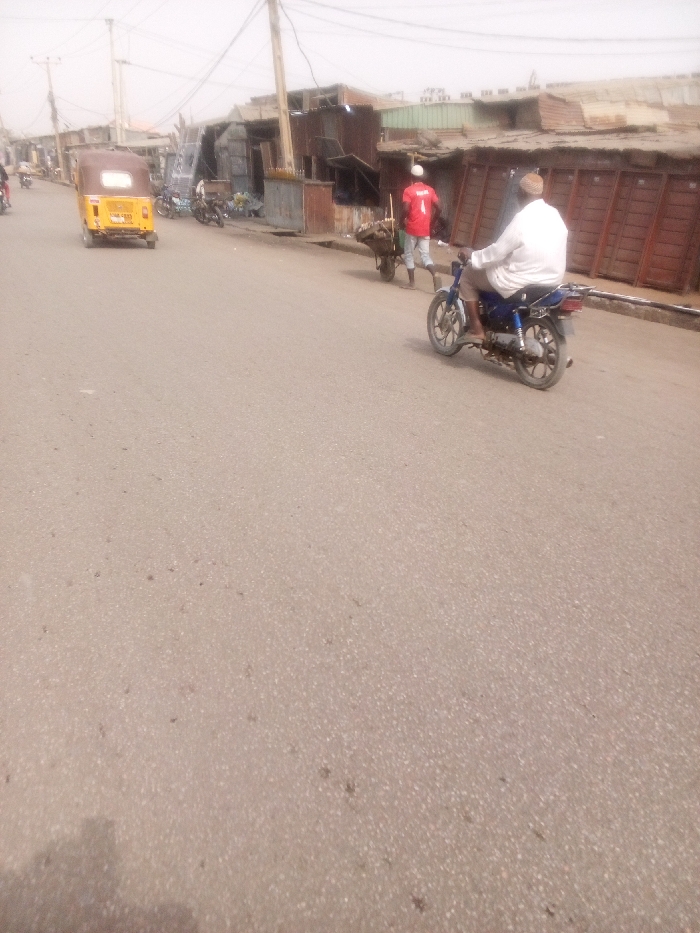 The image captures a daytime street scene, likely in an urban or semi-urban environment, characterized by a hazy or dusty atmosphere. A wide, light-colored asphalt road occupies the foreground and midground of the image.
On the right side of the road in the midground, a man wearing a white shirt or tunic and light-colored trousers is riding a blue motorcycle, moving away from the camera. To his left, another man, dressed in a red t-shirt with the number "1" prominently displayed on the back and a white cap, is pushing a wooden wheelbarrow. The wheelbarrow appears to contain various items. Behind these individuals, a row of informal structures, possibly small shops or stalls, line the roadside. These buildings are constructed predominantly from corrugated metal sheets and wood, with some featuring closed wooden doors or shutters.
On the left side of the road, a yellow three-wheeled auto-rickshaw, seen from its rear, is also moving away from the camera. Further down this side of the street, other vehicles, possibly motorcycles or more auto-rickshaws, are visible, either parked or in transit. Similar makeshift structures also line this side of the road.
The background is somewhat blurred and indistinct due to the hazy conditions, revealing more buildings and numerous utility poles with a dense network of overhead electrical or communication wires, extending into the distance. The sky is bright but appears overcast or dusty, consistent with the overall hazy quality of the image. The scene depicts typical daily activity on a community street.