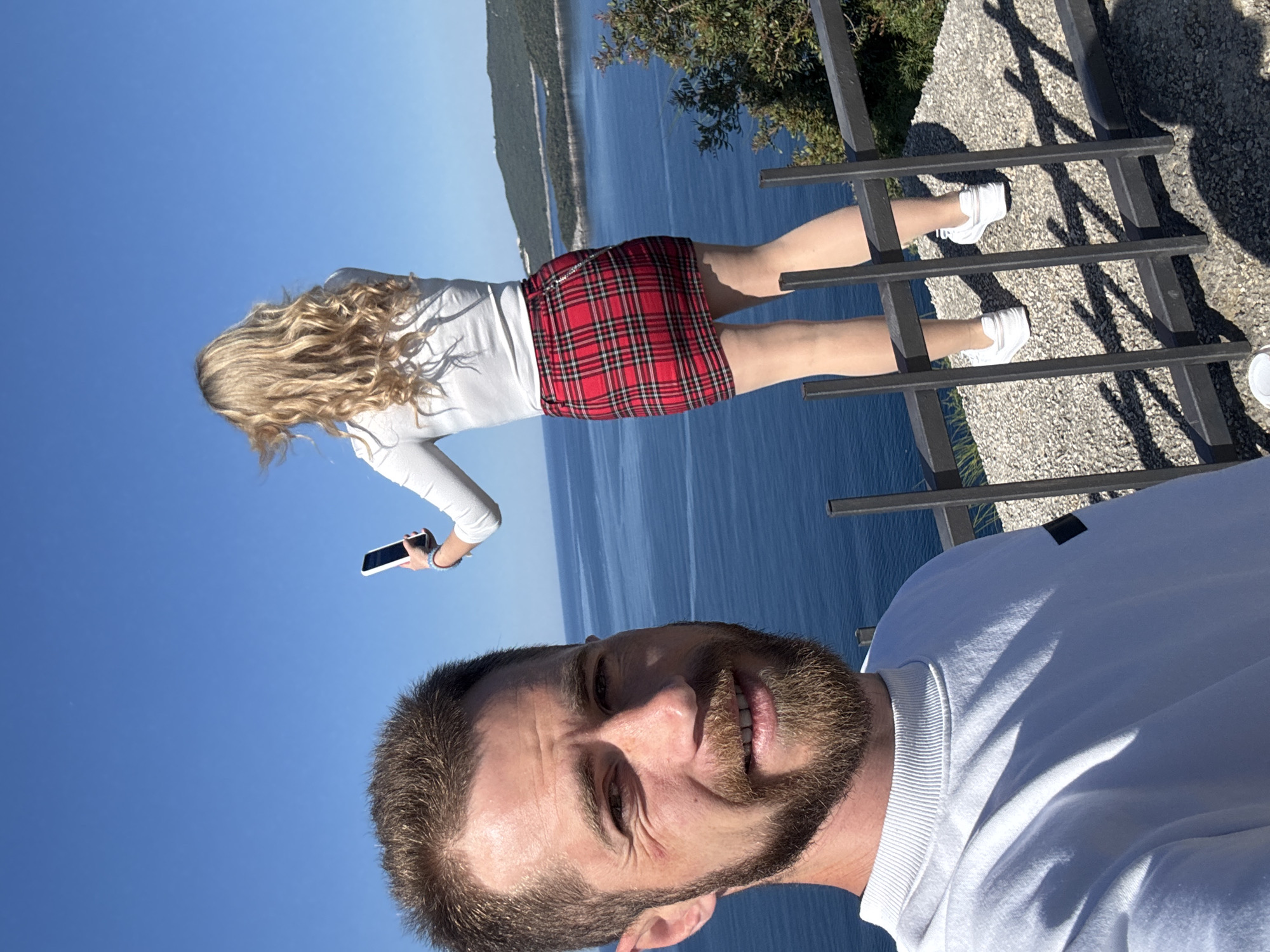 A selfie featuring a man with a beard, wearing a white sweatshirt, is oriented vertically. Behind him, a woman in a white long-sleeved shirt and red plaid skirt stands on metal railings, holding a smartphone. The background shows a body of water with distant land, suggesting a coastal location. The location is likely near Budva, Montenegro, given the typical coastal landscapes found there. A patch of greenery is visible in the upper right corner, possibly indicating a nearby bush or tree.