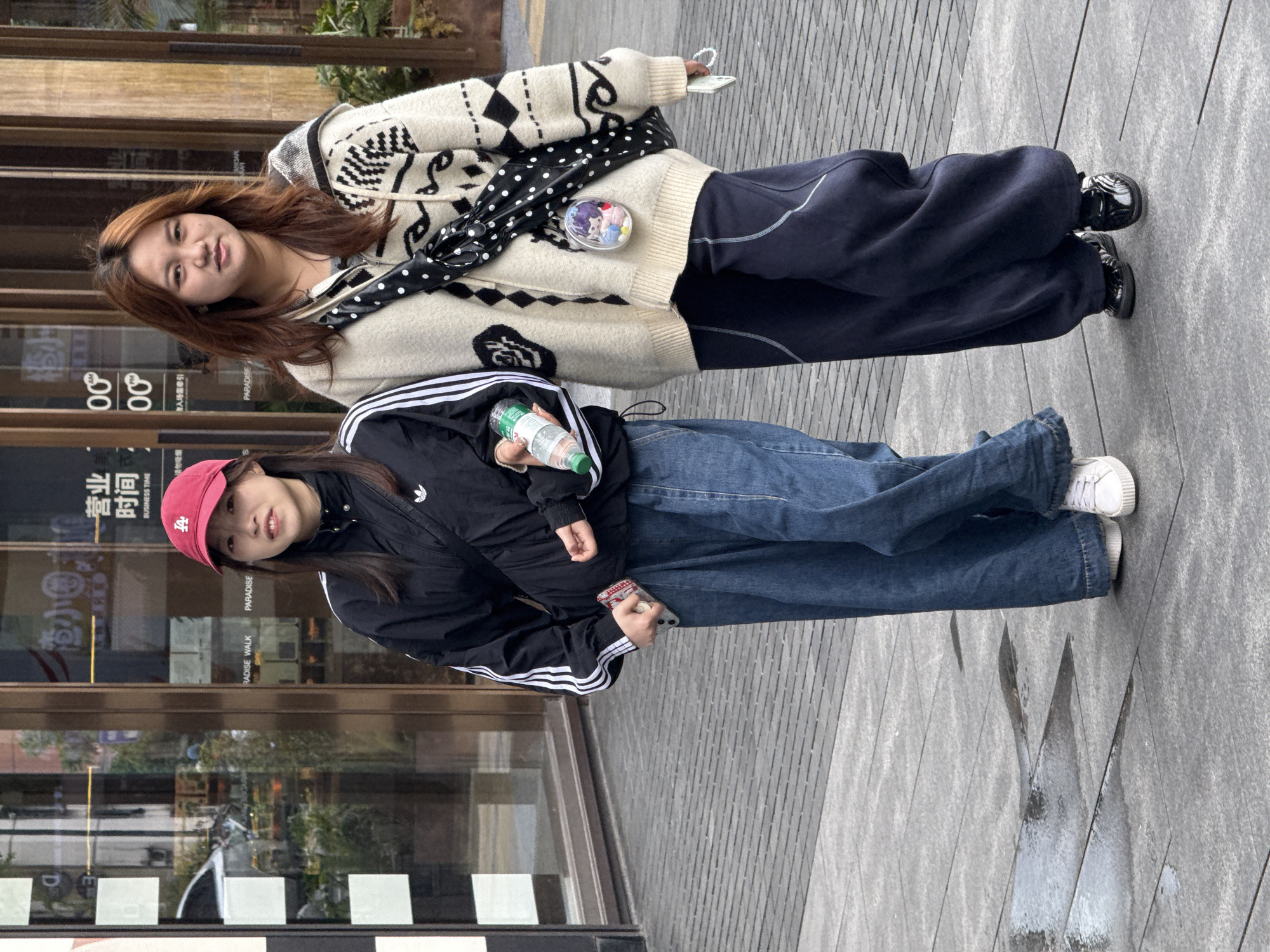 Two young women stand side-by-side on a gray, textured walkway in what appears to be an urban area. The woman on the left wears a black Adidas jacket with white stripes, a red baseball cap featuring the letter "A," and blue jeans. She holds a water bottle in her right hand and a mobile phone in her left hand. The woman on the right wears a cream-colored cardigan sweater with black patterns, navy blue pants, and carries a black purse with white polka dots. The background shows a storefront with text in both Chinese and English, including "BUSINESS TIME," "PARADISE WALK," and "PARADISE." The location is identified as being in Yuzhong District, China.