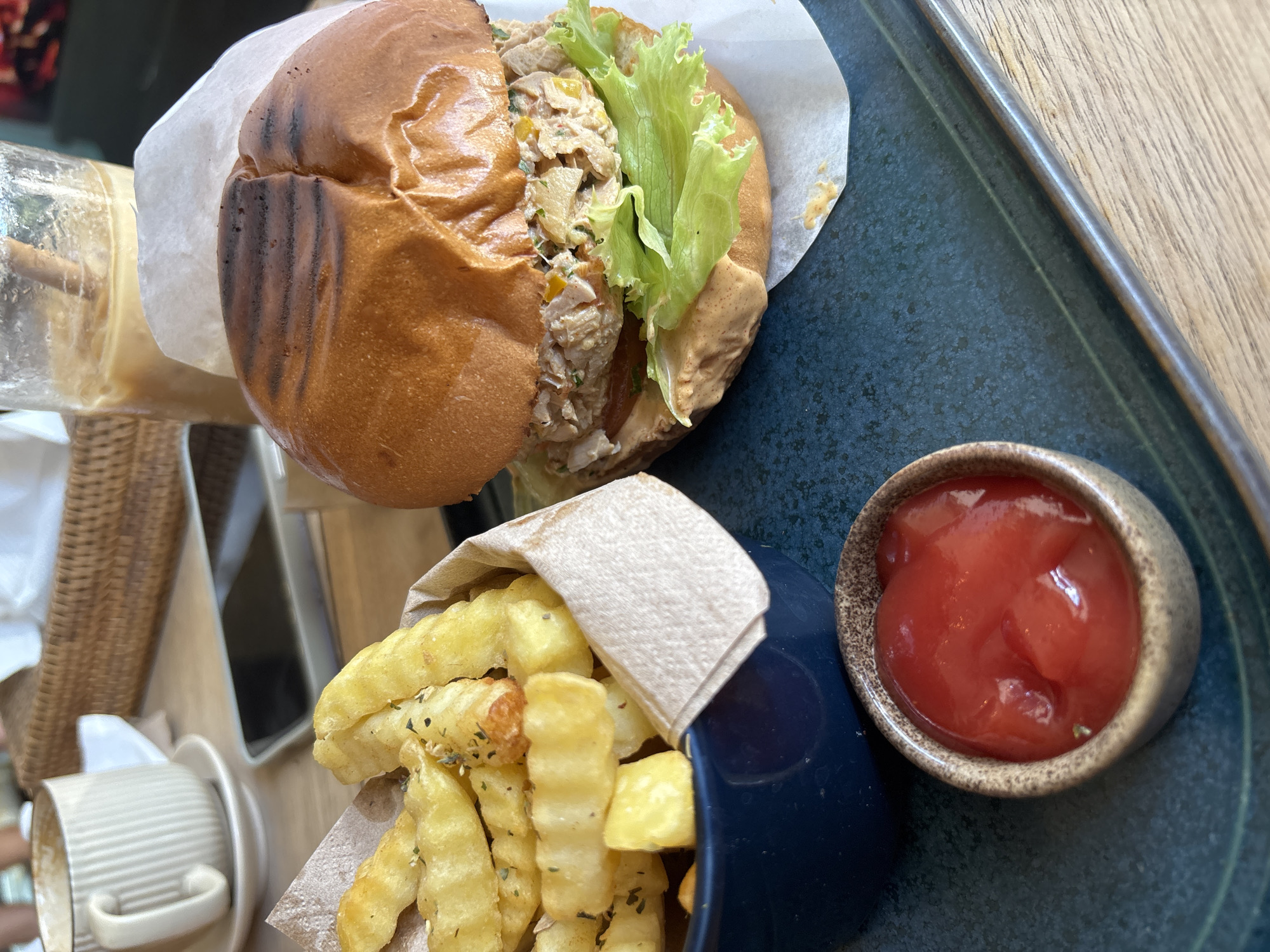 On a wooden table in Kuta Selatan, Indonesia, a dark blue tray holds a chicken salad sandwich on a white paper liner, served with French fries in a blue container with a paper lining, and a small bowl of ketchup. The sandwich is comprised of a toasted bun, chicken salad filling, lettuce, and sauce. A glass of liquid is partially visible next to the sandwich. A cup and saucer are situated near the French fries. The scene is photographed from an overhead perspective.