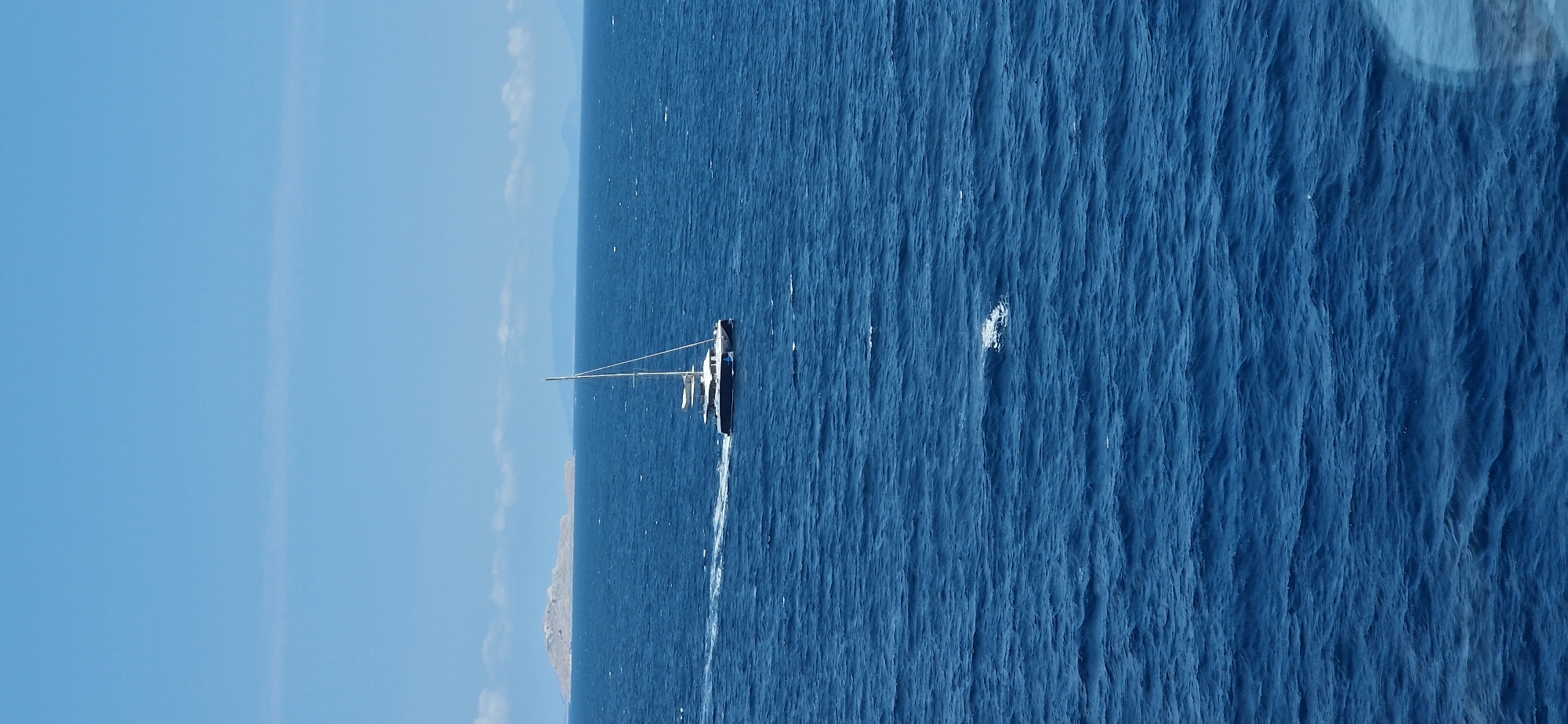 A catamaran sailboat with a single mast is sailing on a deep blue sea. The boat is white with a dark hull and is leaving a white wake behind it. In the distance, there is a small, rocky island. The sky is clear and blue with a few wispy clouds. The scene appears to be set in the Municipal Unit of Agioi Theodoroi, Greece, given the characteristic Mediterranean island and clear blue sea.