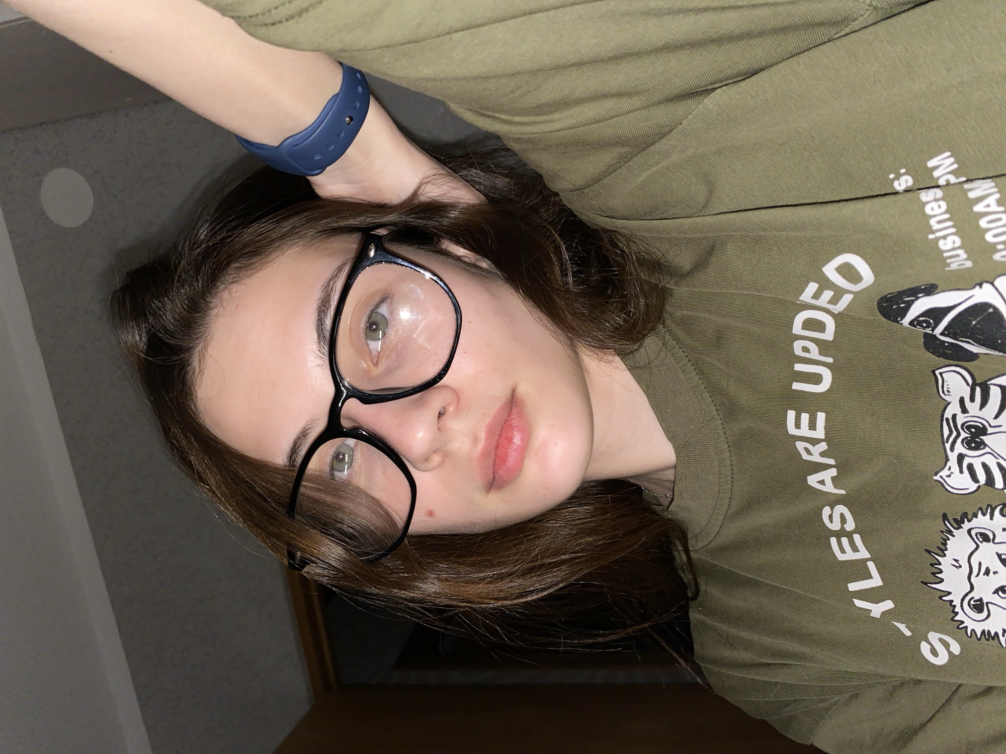 The image shows a young woman wearing glasses, a blue wristband, and an olive green shirt with white text and animal graphics. She is holding her head with her arm, and the photo appears to be taken indoors. It is impossible to determine if the location is in Votkinsk, Russia from the image provided.