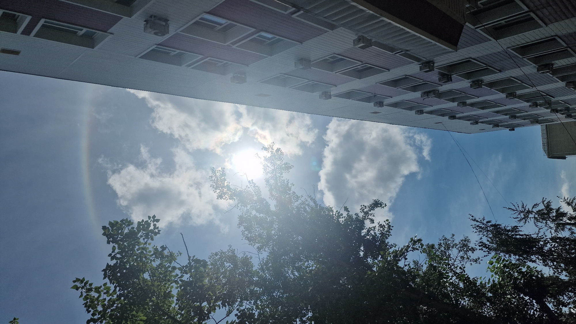 The image shows a view looking upwards towards the sky and a building. On the left, the branches and leaves of a large tree are visible against a bright blue sky with scattered white clouds. The sun is partially obscured by clouds, creating a lens flare effect and a faint rainbow or sun halo is visible in the upper part of the sky. On the right, the side of a modern, multi-story building is prominent, with its facade featuring a repeating pattern of windows and maroon-colored accents. Several air conditioning units are mounted on the building's exterior. The building appears to be located in Kostanay, Kazakhstan. The overall impression is that of an outdoor urban scene on a sunny day.
